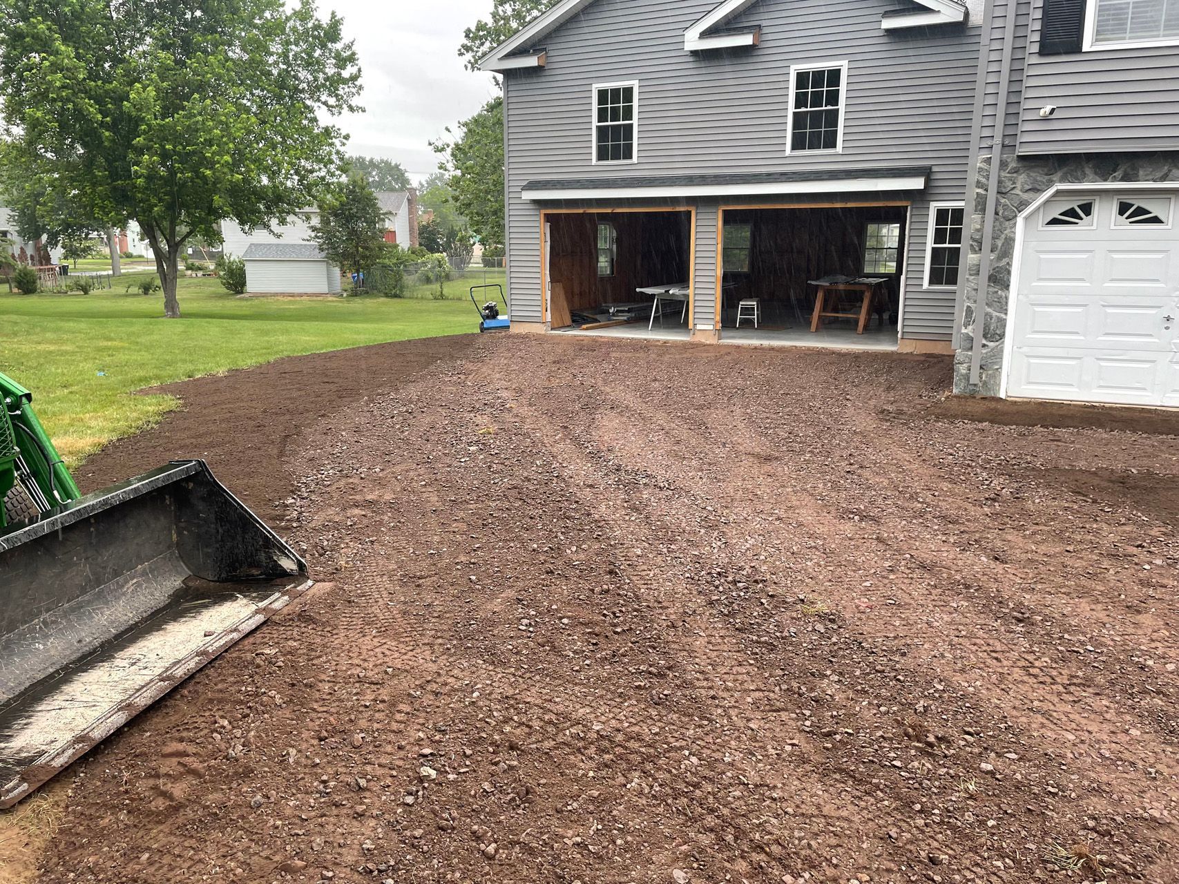Freshly spread gravel driveway in front of a gray house with a white garage door and open carport, with a skid-steer bucket at left