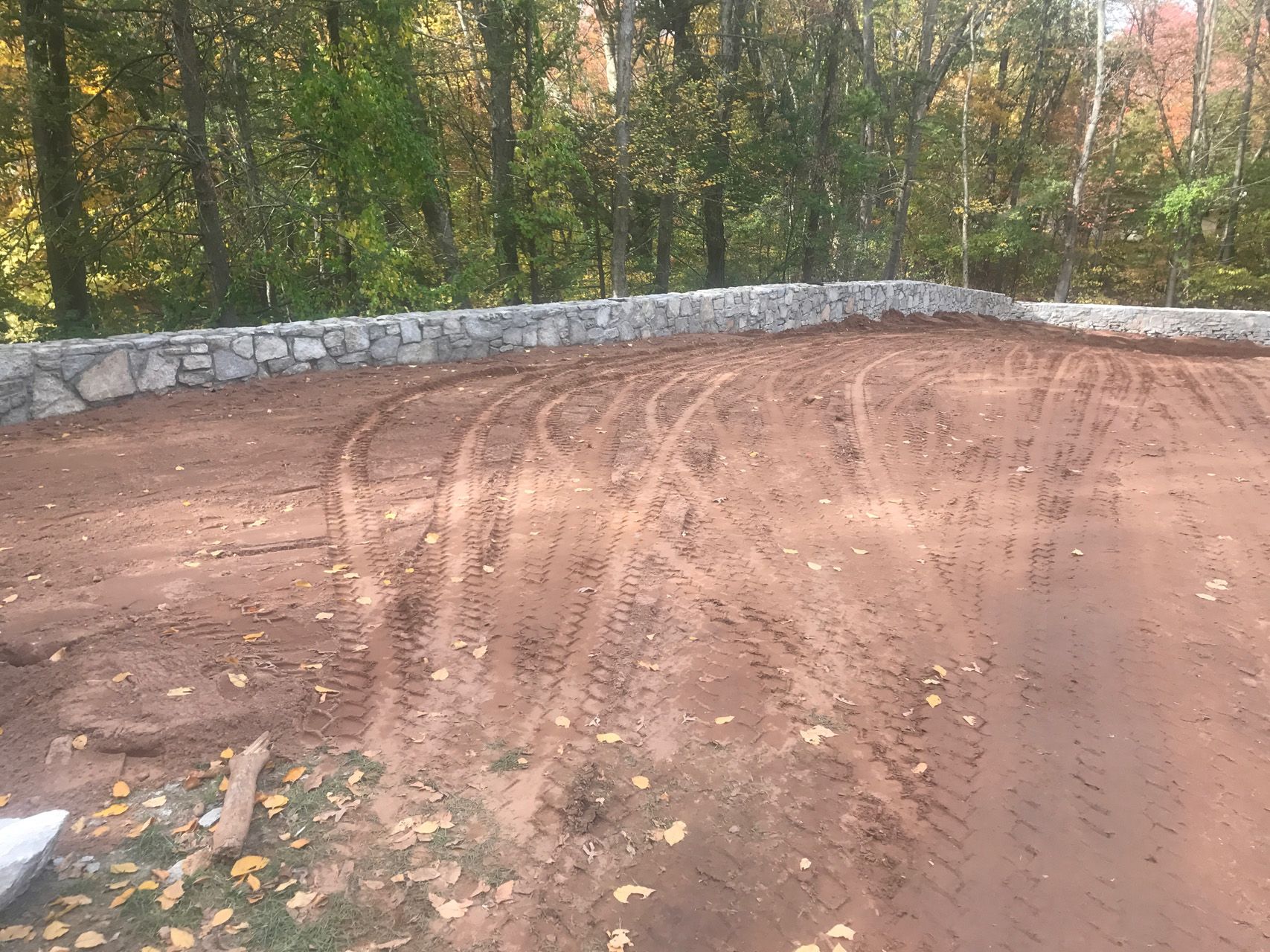 Freshly graded dirt lot with tire tracks beside a stone wall and trees in the background