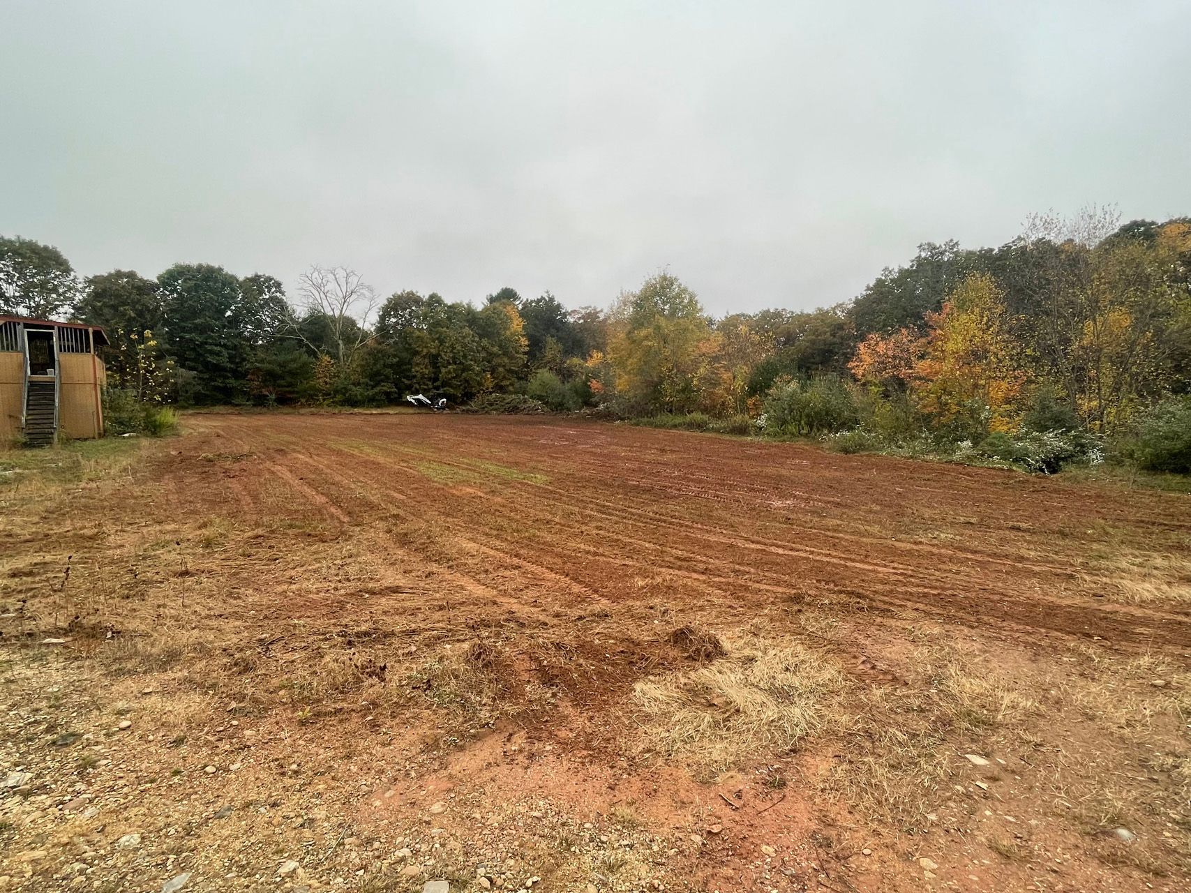 A freshly cleared brown field with trees showing autumn colors under a cloudy sky.