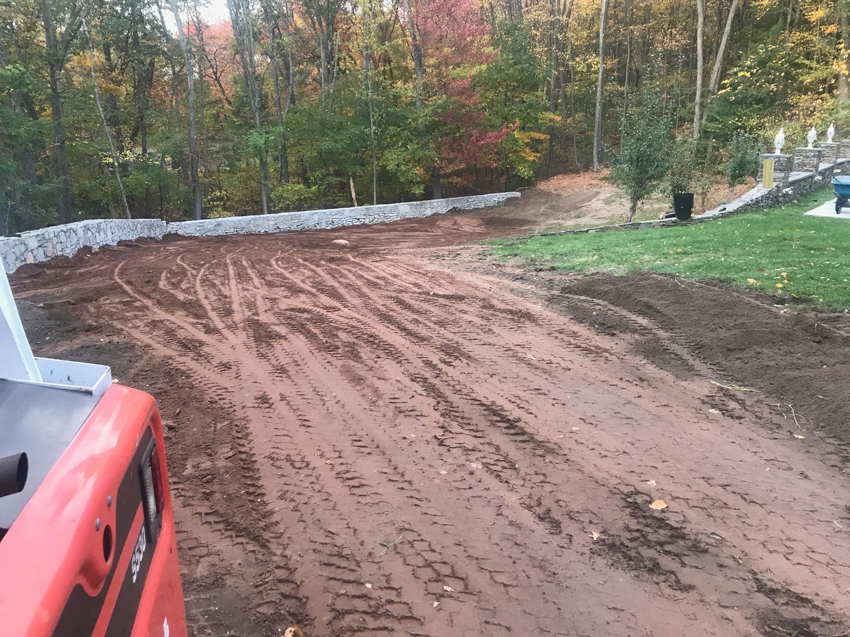 Freshly graded dirt driveway beside a white retaining wall, with a red vehicle edge and autumn trees beyond.