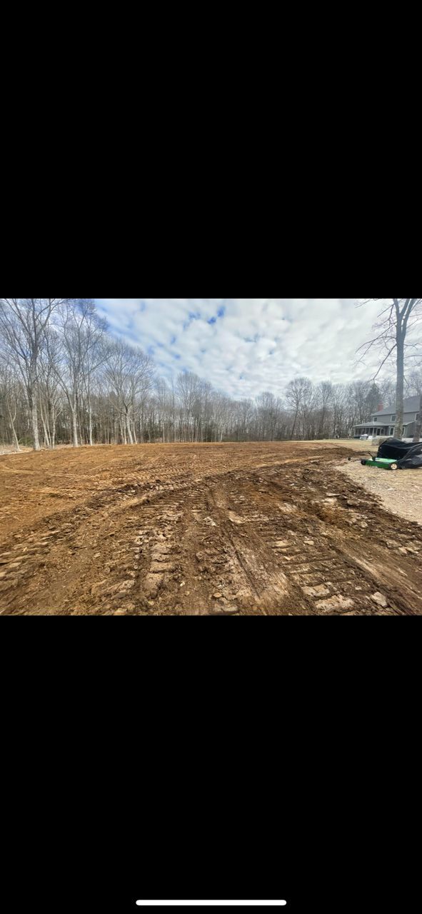 Dirt lot with tire tracks beside leafless trees under a cloudy sky, with a parked vehicle at right.