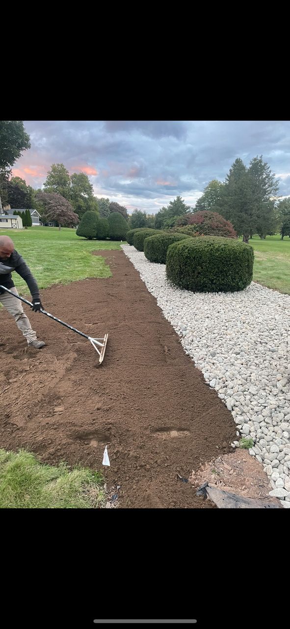 Worker spreading soil beside a white gravel path in a landscaped yard under cloudy skies