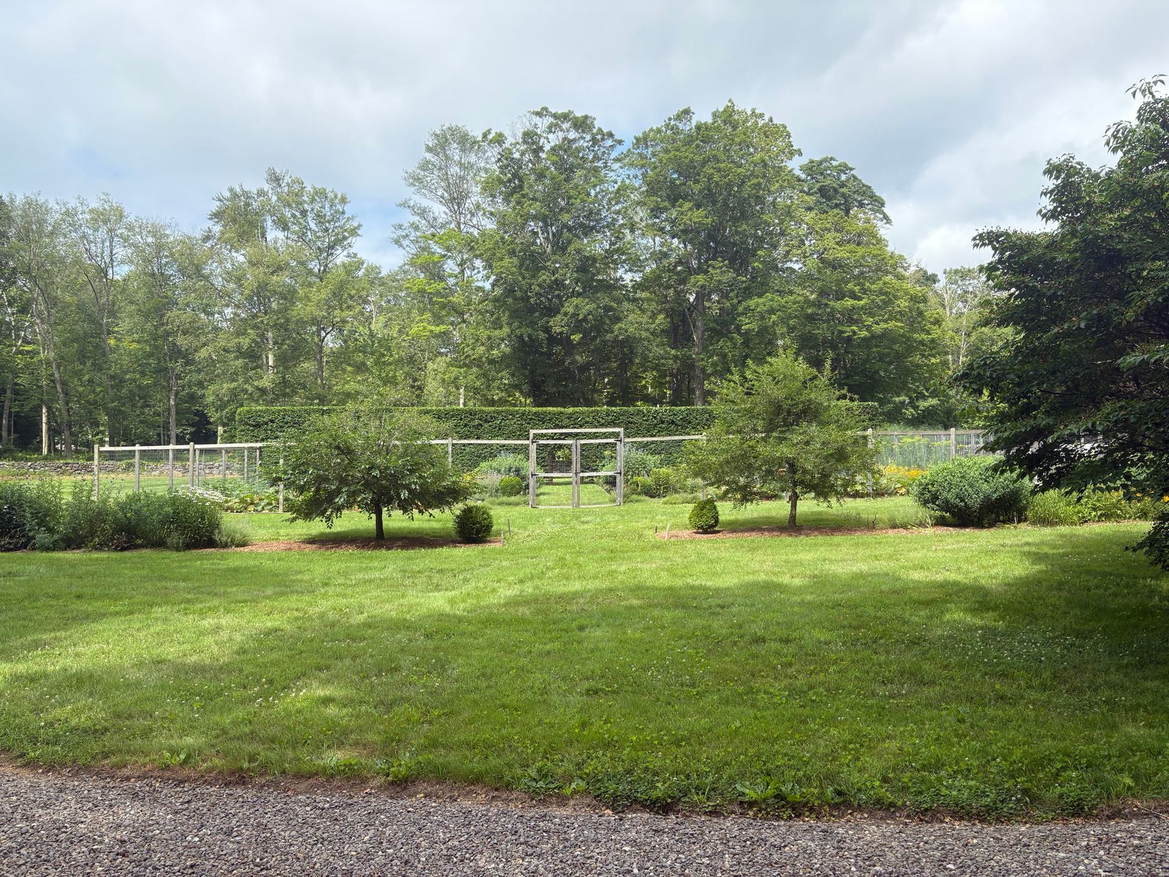 Grassy park with trees, shrubs, and a white fence under a cloudy sky
