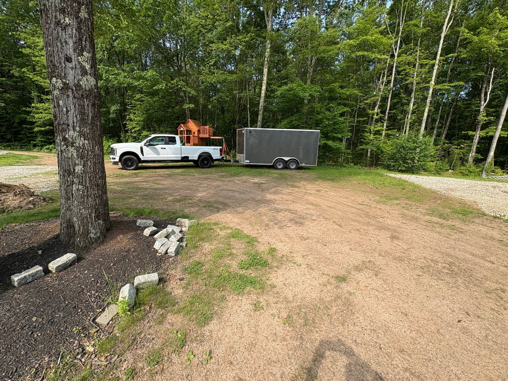 White pickup truck hitched to a gray trailer parked on a dirt driveway in a wooded area.