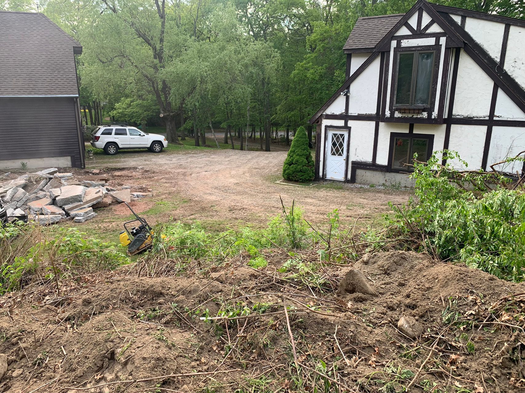 Rural yard with dirt mound, old car, and a white-and-black Tudor-style house by trees