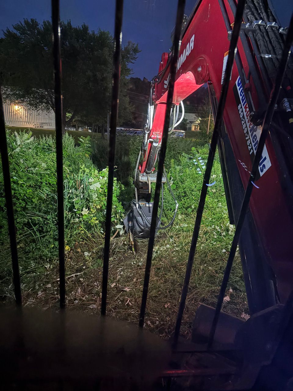 Red excavator arm working behind black metal fence beside bushes at night