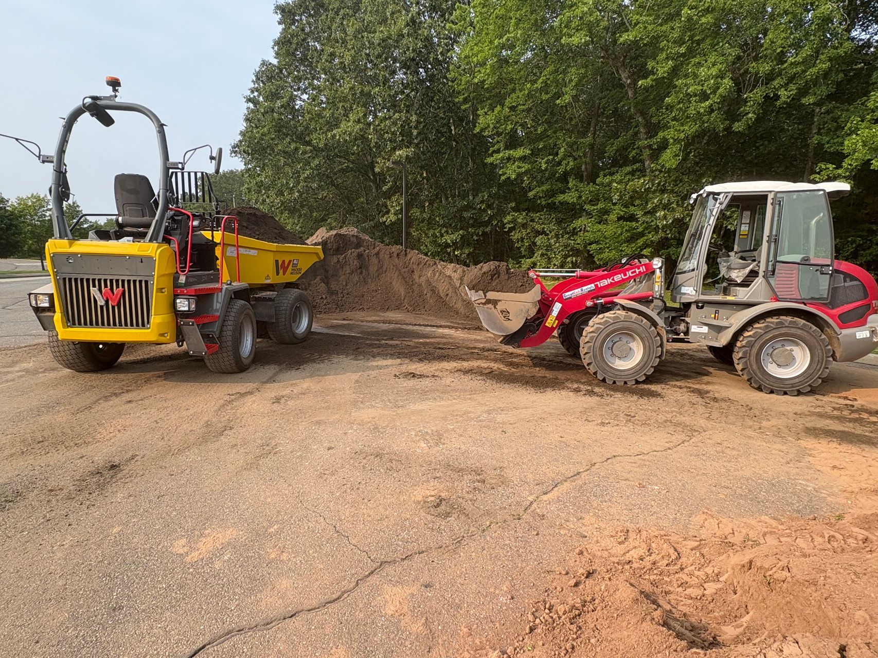 Yellow dump truck and red loader on a dirt road near trees and a mound of soil