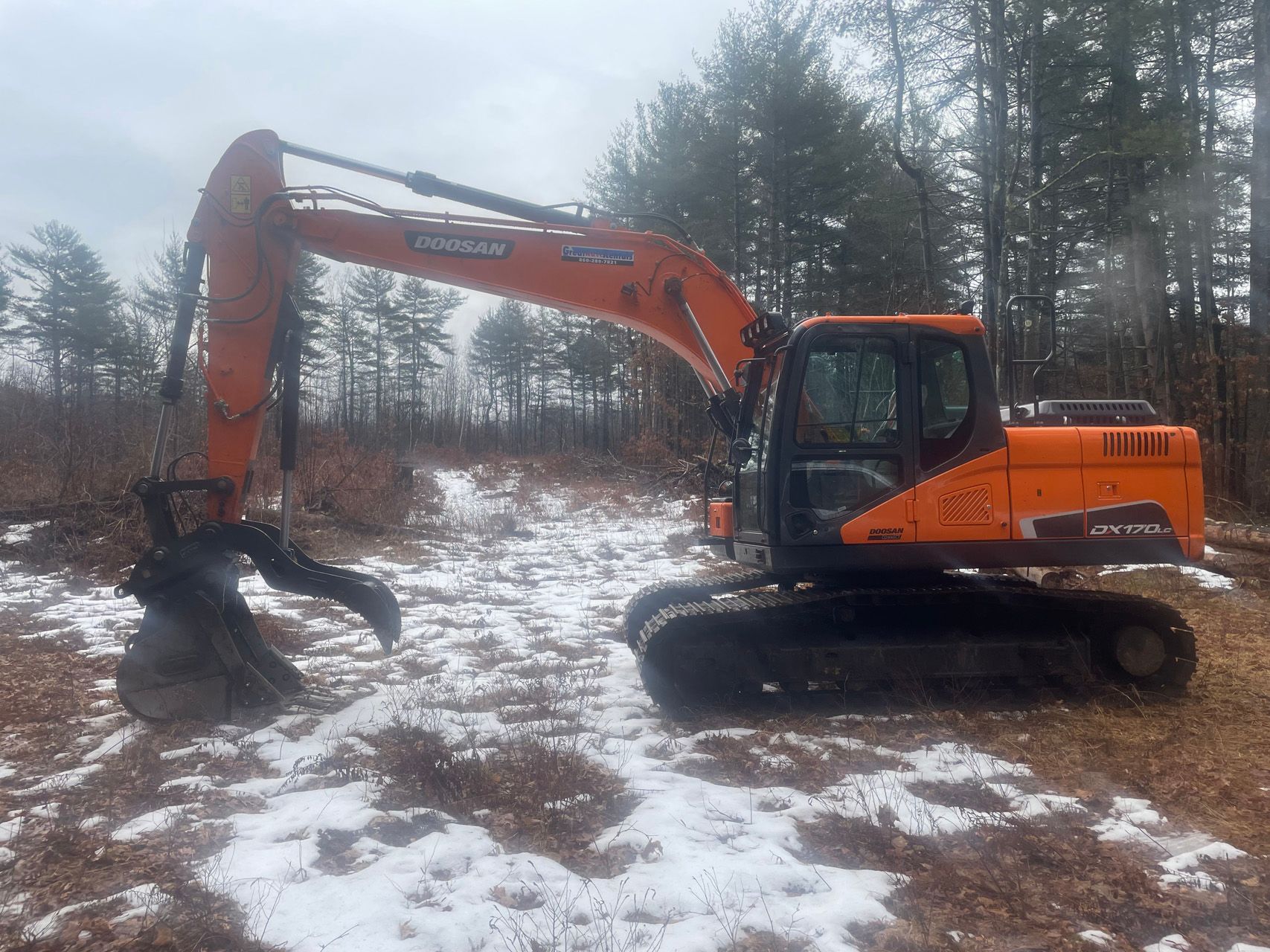 Orange excavator on snowy ground in a wooded area, with its bucket lowered.