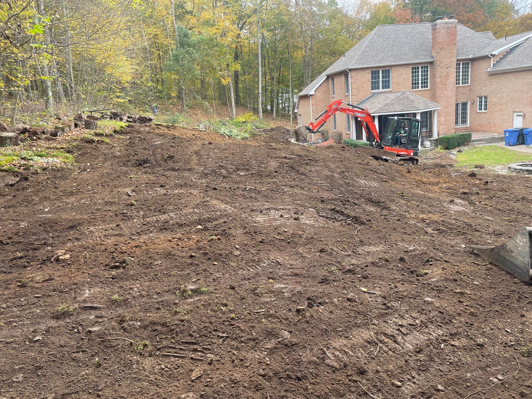 Freshly graded dirt lot beside a brick house, with a red excavator and autumn trees in the background