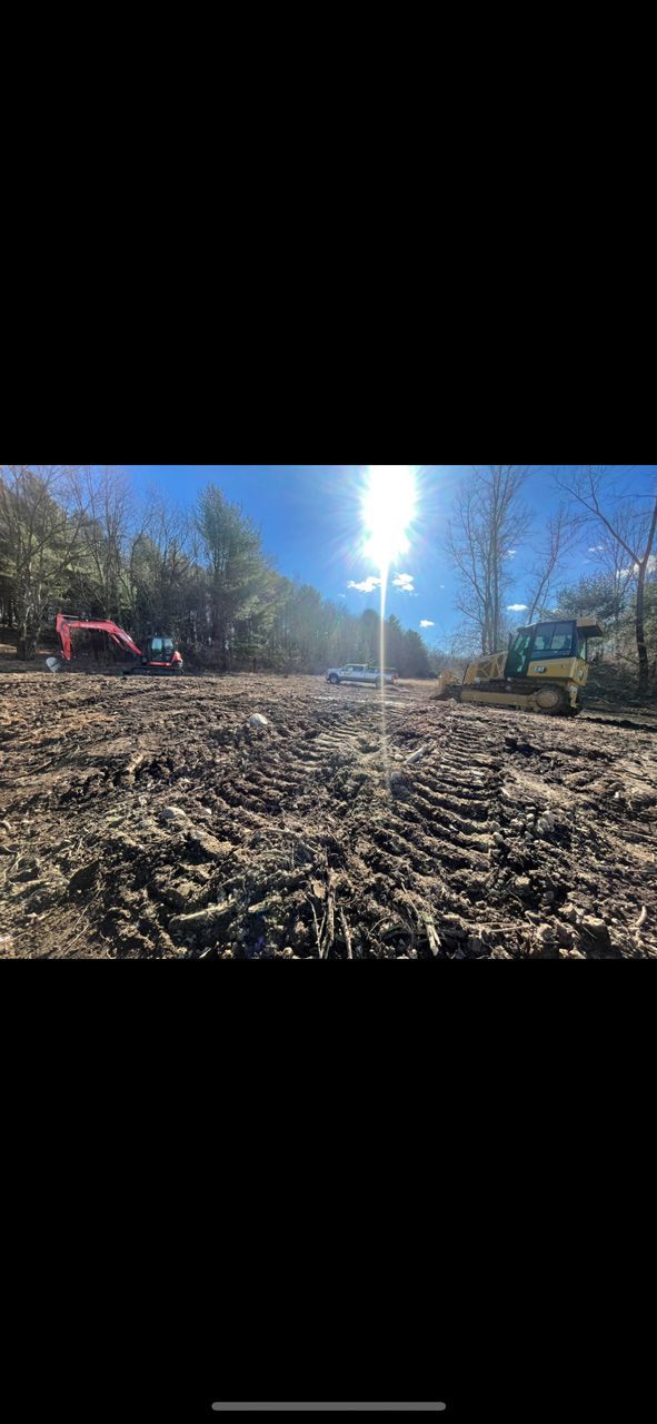 Sunlit dirt lot with wood chips, trees, a pink excavator, and a small building in the background