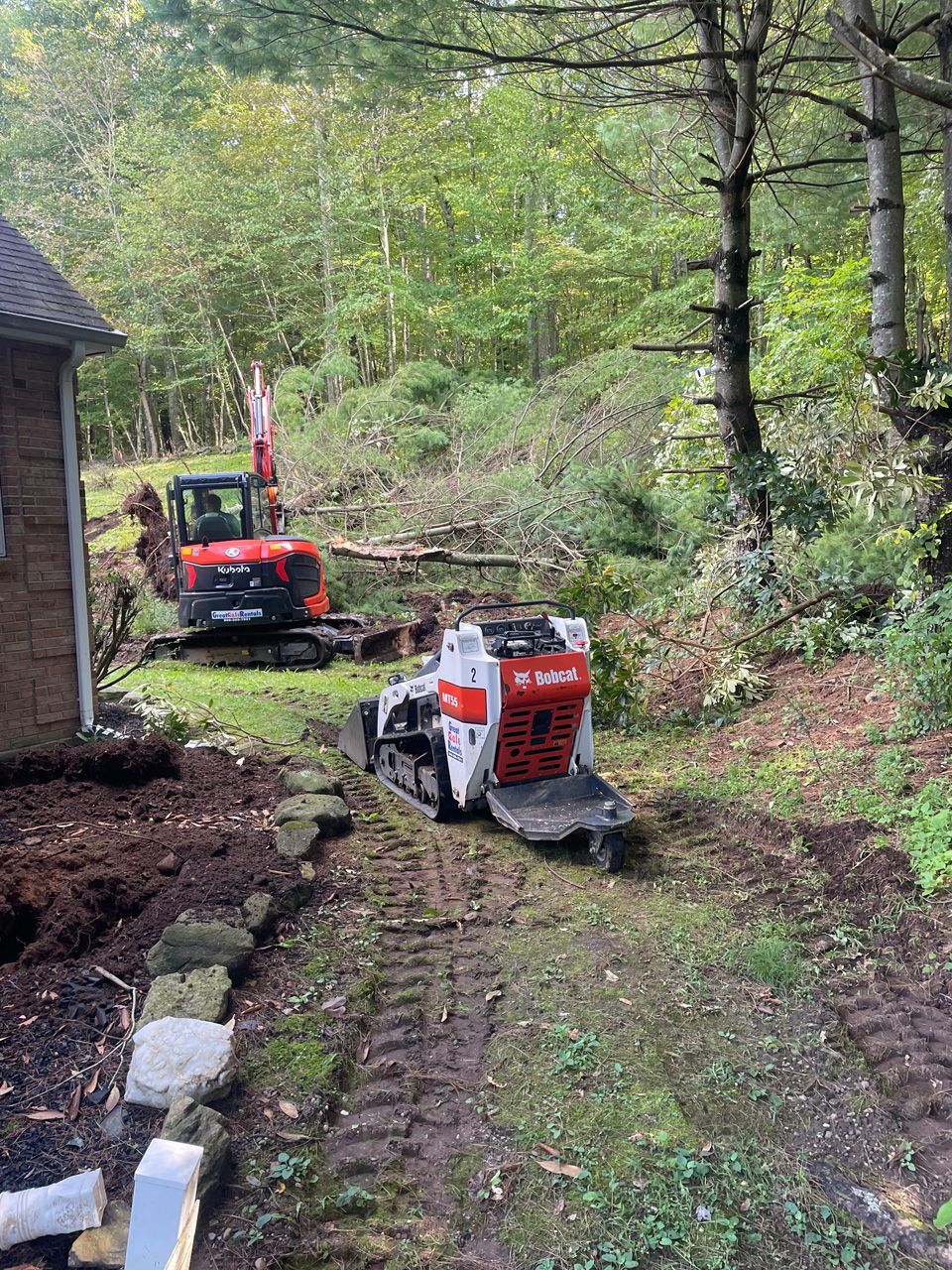 Two construction machines on a muddy wooded path beside a house, clearing debris.