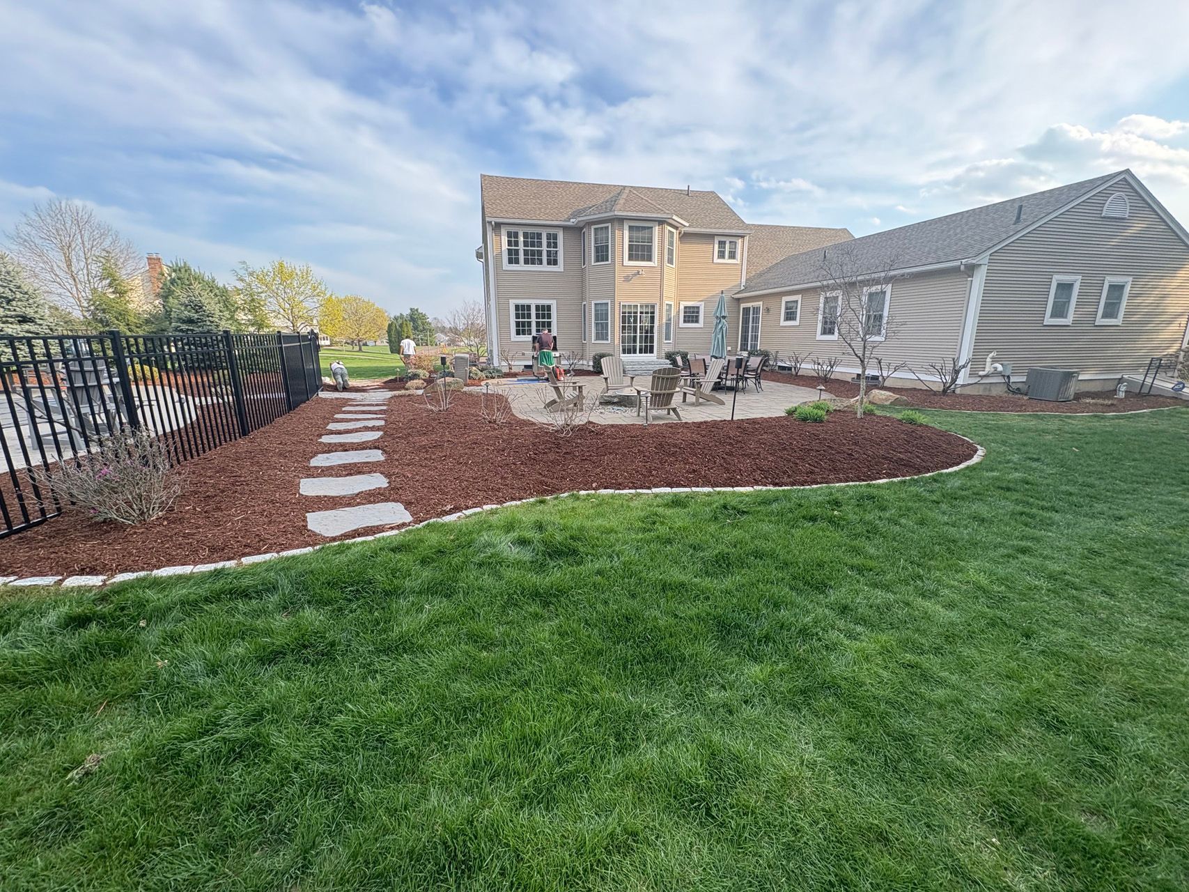 Backyard patio with stone path, red mulch beds, trimmed lawn, and a two-story house under a cloudy sky