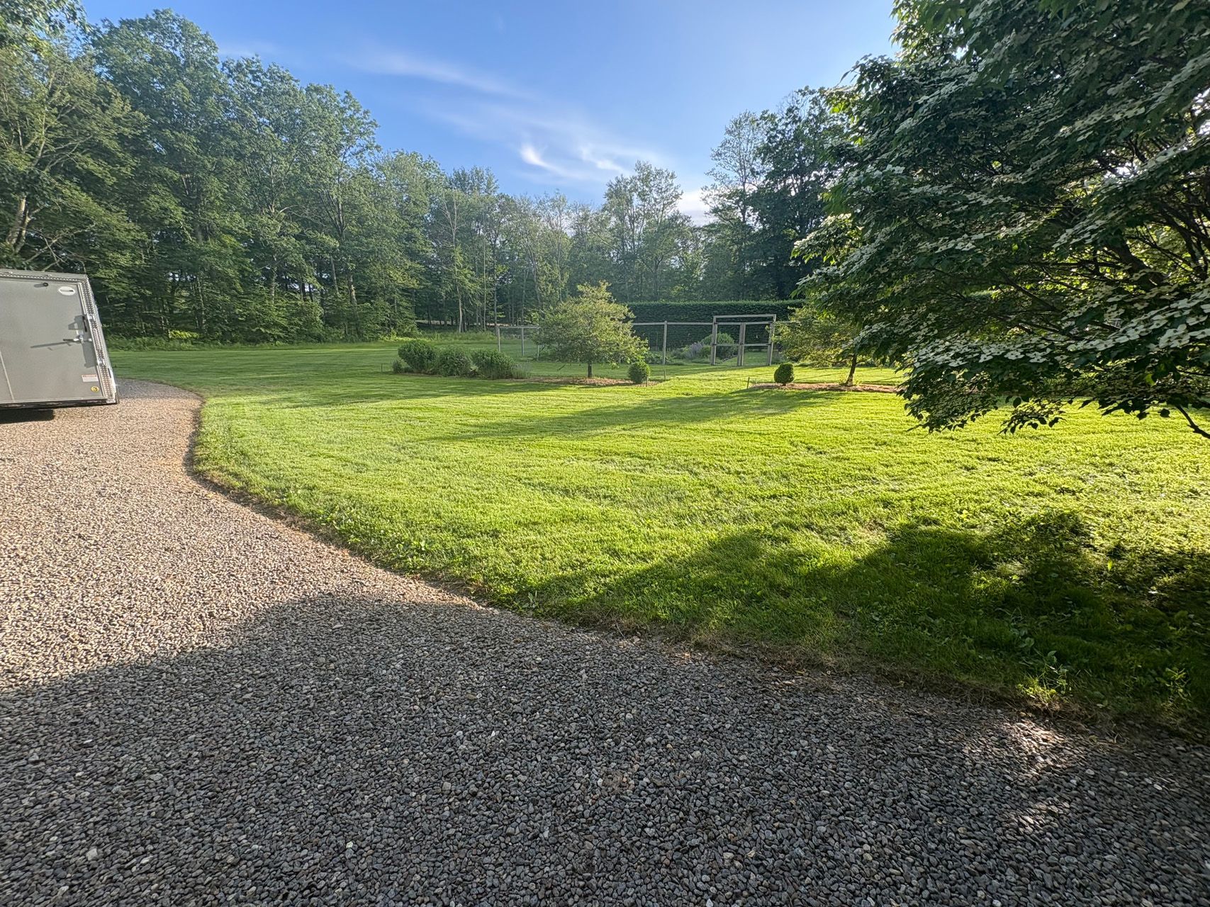 Sunlit gravel driveway beside a green lawn and trees, with a trailer at left and a fenced area in back