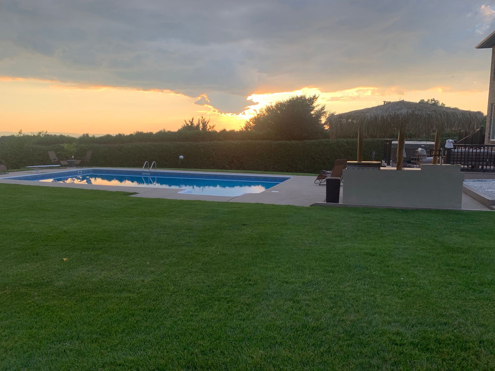 Outdoor pool area at sunset with a green lawn, blue covered pool, and deck chairs under cloudy sky