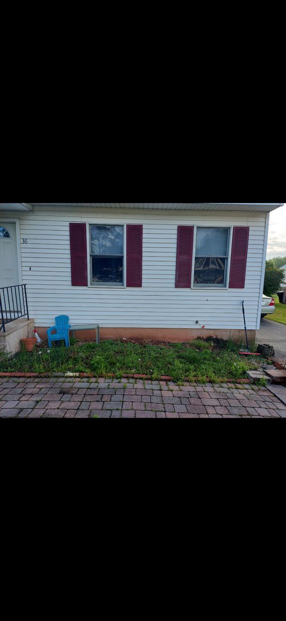 Small white house exterior with red shutters, a blue bucket, and a brick walkway in front.