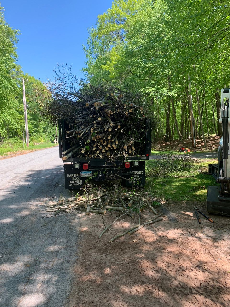 Trailer piled high with branches on a rural road beside green trees and sunlight
