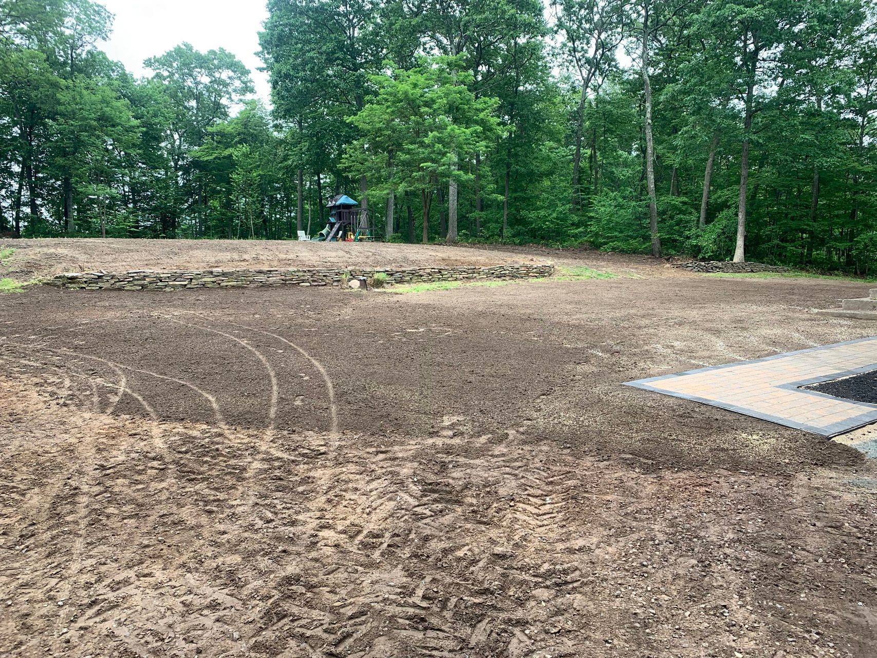 Cleared dirt lot beside trees with a small worker in the distance and a concrete slab at right