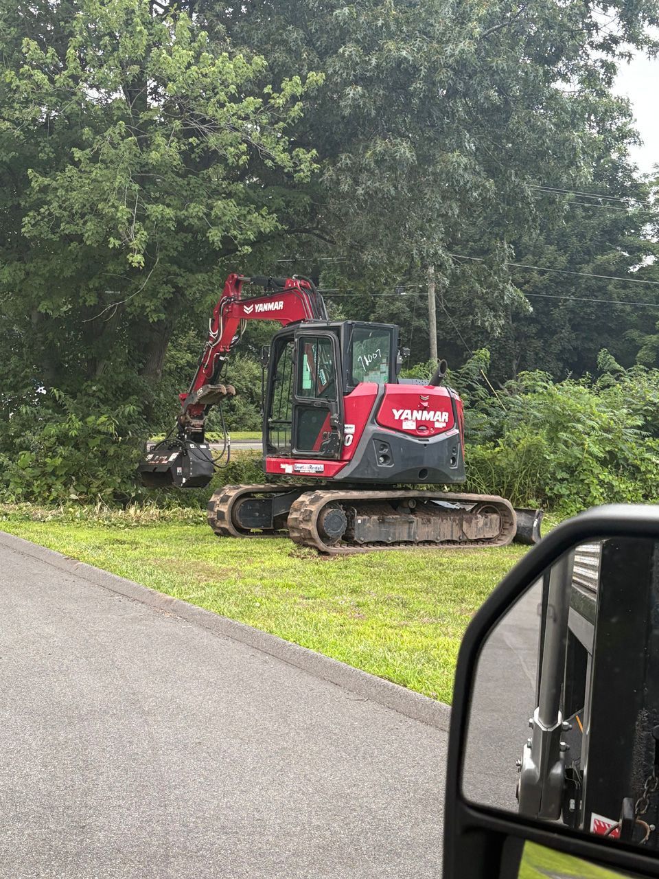 Red excavator parked beside a road at the edge of a grassy, wooded area.