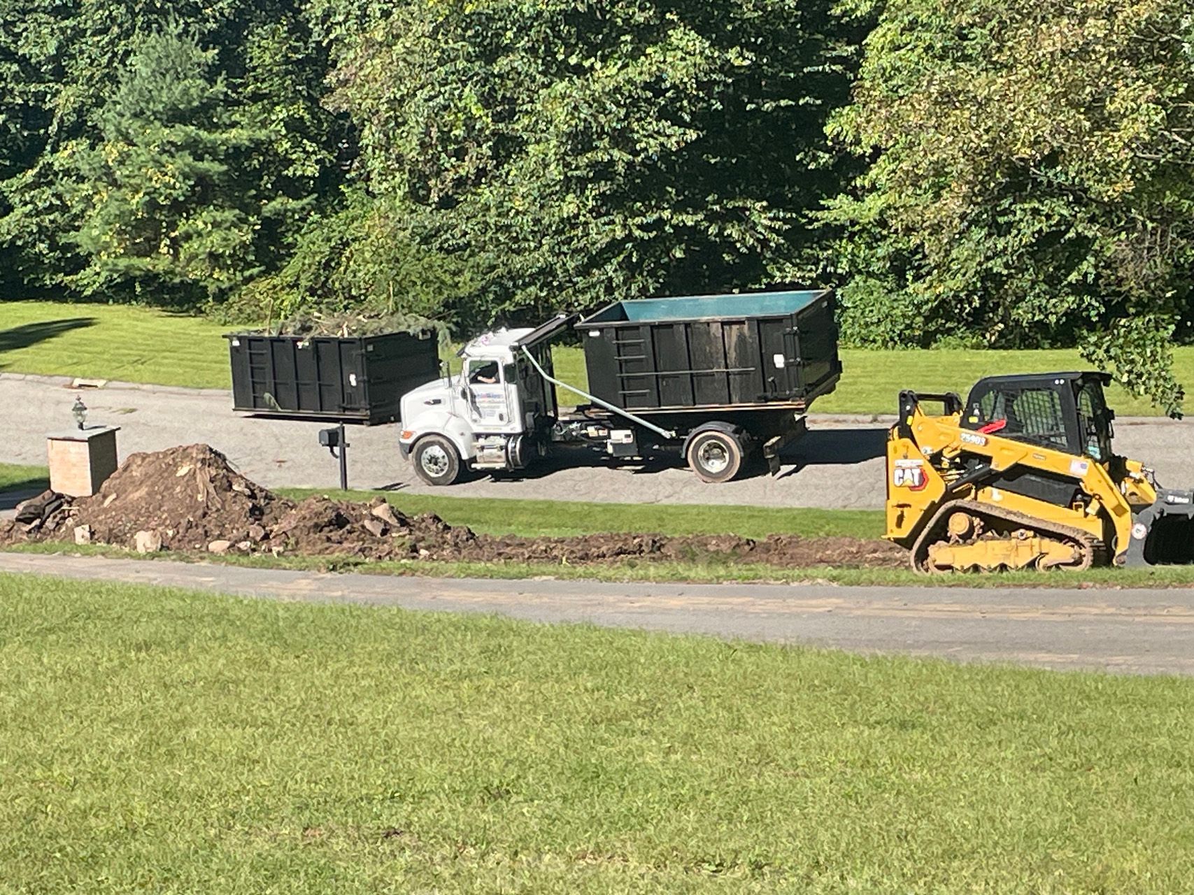 Construction equipment and dump truck beside a dirt pile on a grassy roadside near trees