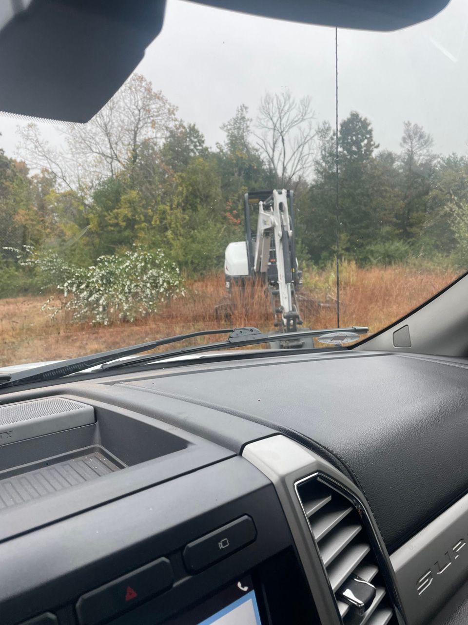 Car interior view of a small loader or excavator ahead on a dirt road with trees and brush in the background