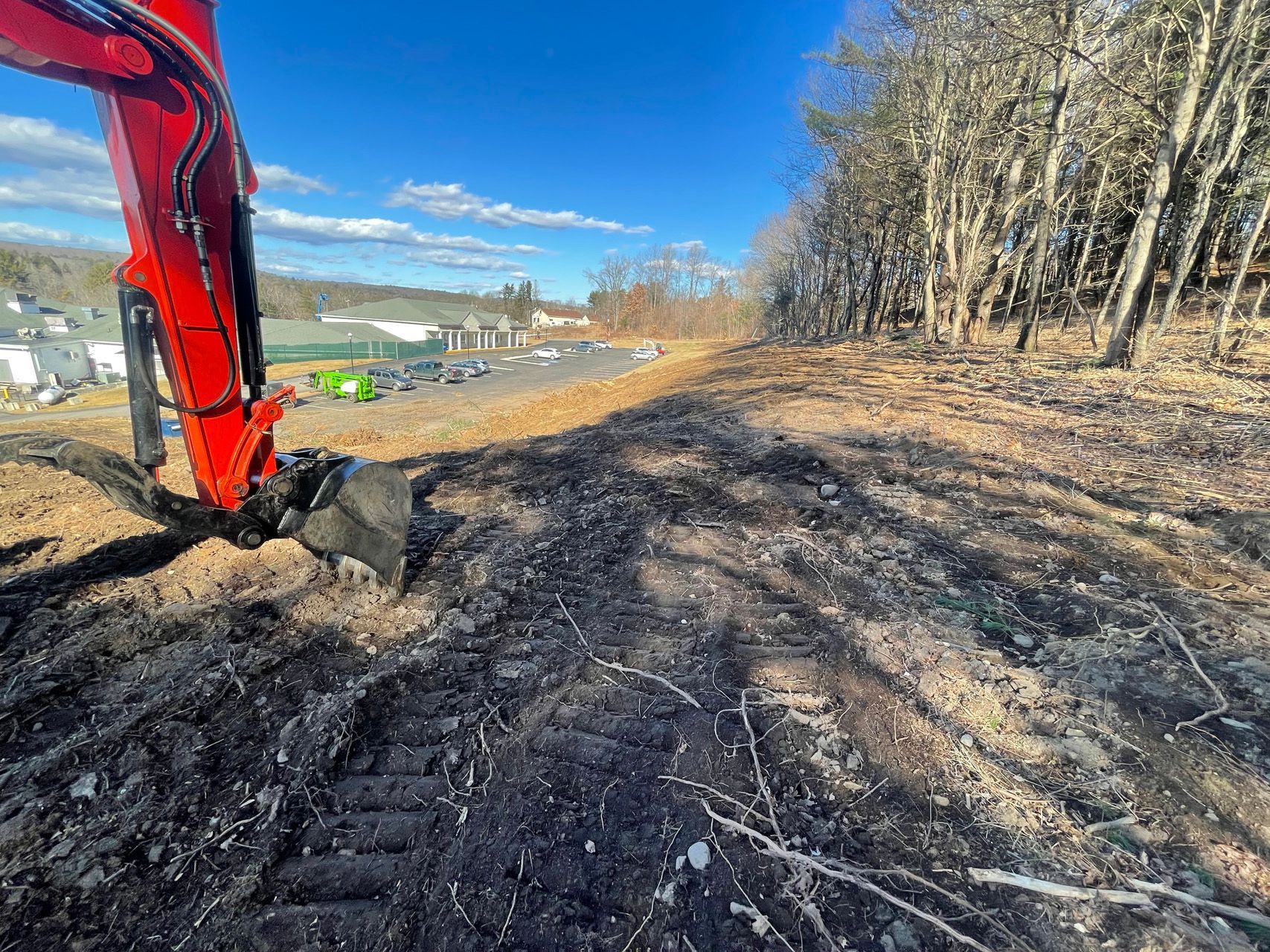 Red excavator digging beside a dirt road under a blue sky, with trees and construction work in the background