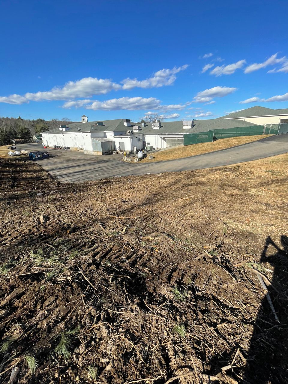 Rural farm buildings beside a dirt road under a bright blue sky, with plowed fields in the foreground