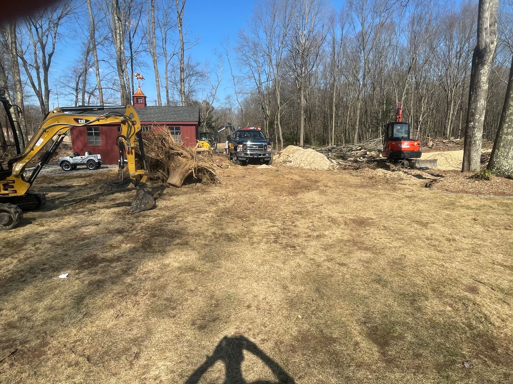 Cleared wooded lot with excavators, trucks, and a red building under a blue sky