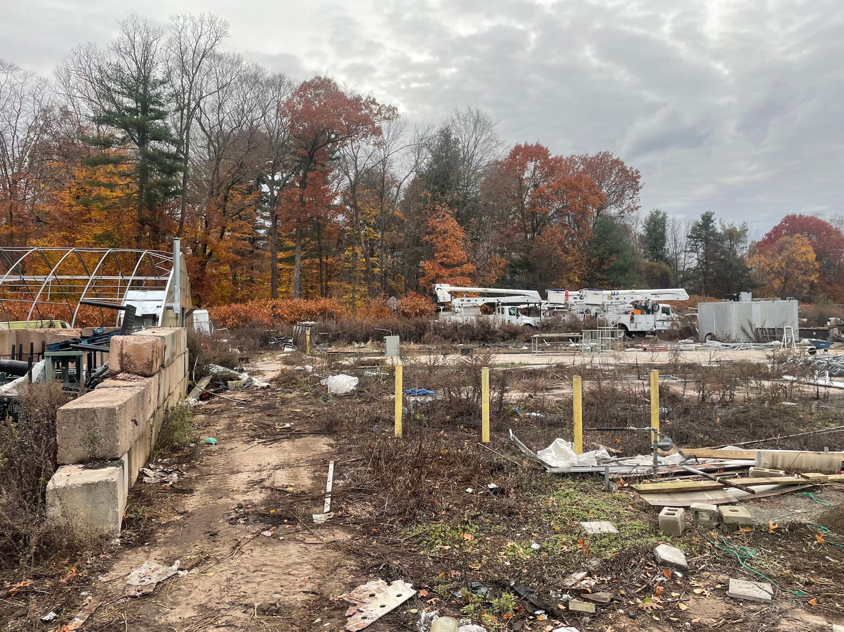 Construction site with concrete foundation, yellow posts, debris, and autumn trees under cloudy sky