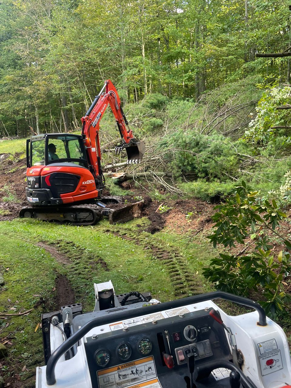 Orange excavator clearing dirt on a wooded hillside, viewed from inside another machine.