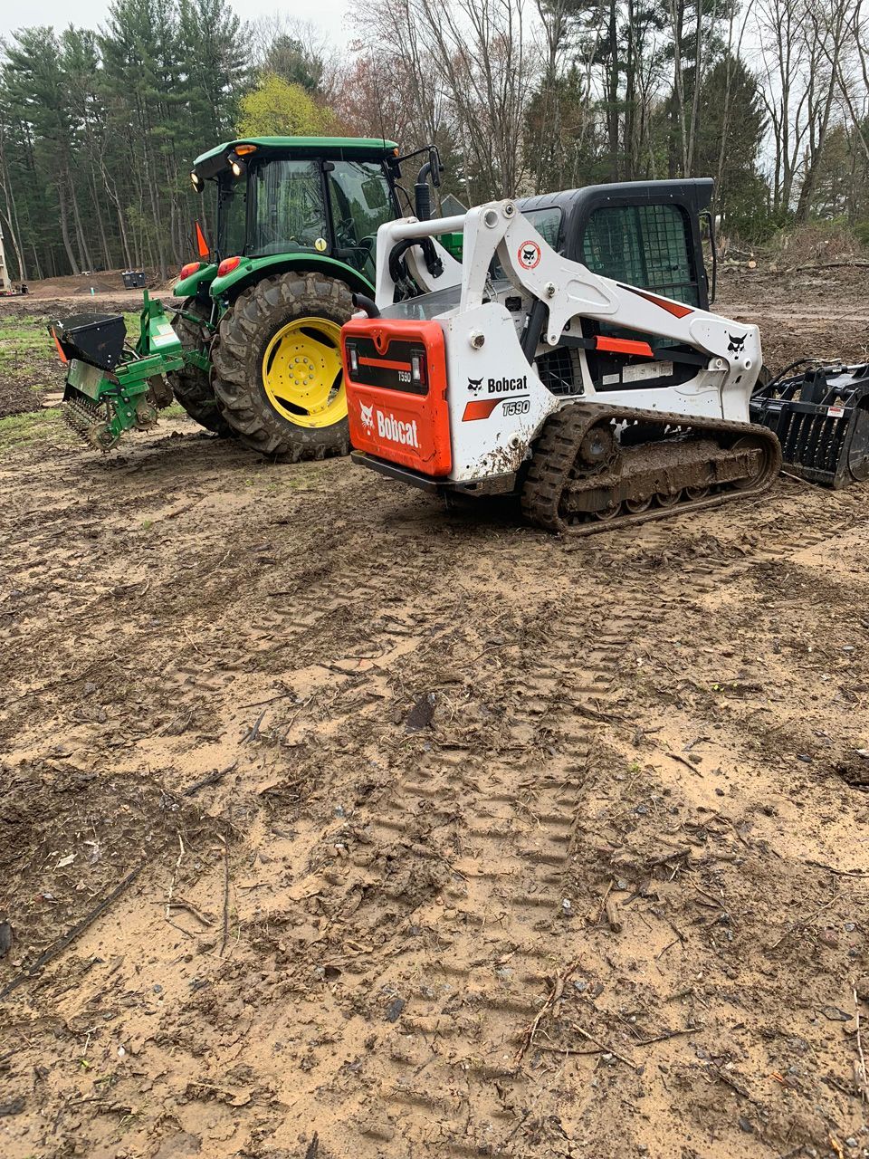 Green tractor and white skid steer on muddy ground in a wooded field.