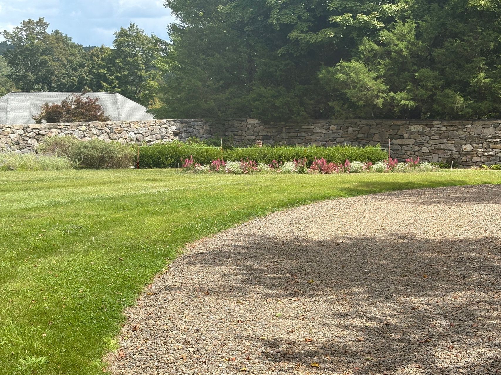 Curved gravel path beside a green lawn, stone wall, and flower bed in a sunny garden