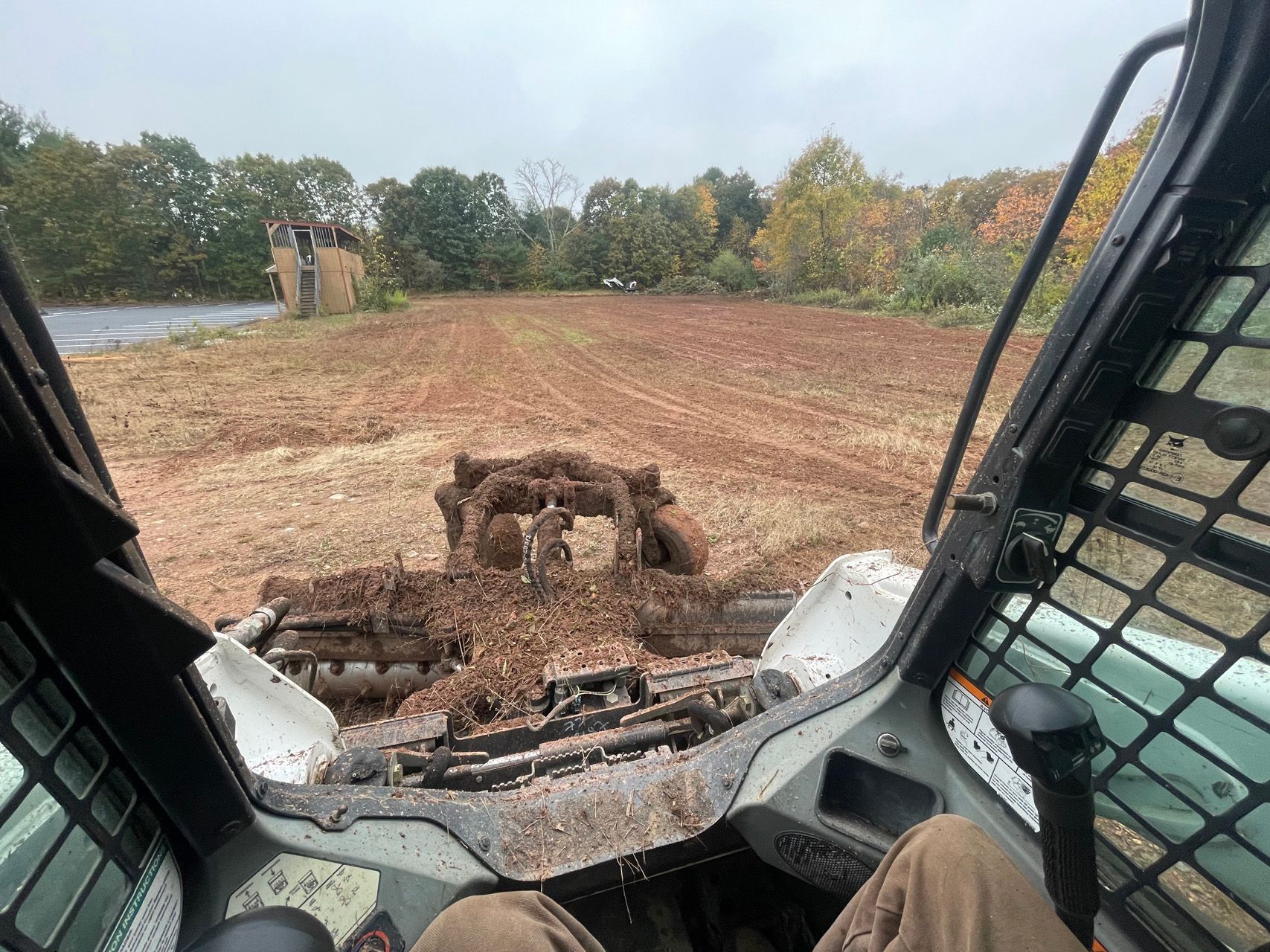 View from a skid steer cab, clearing brown dirt on a wooded construction site.