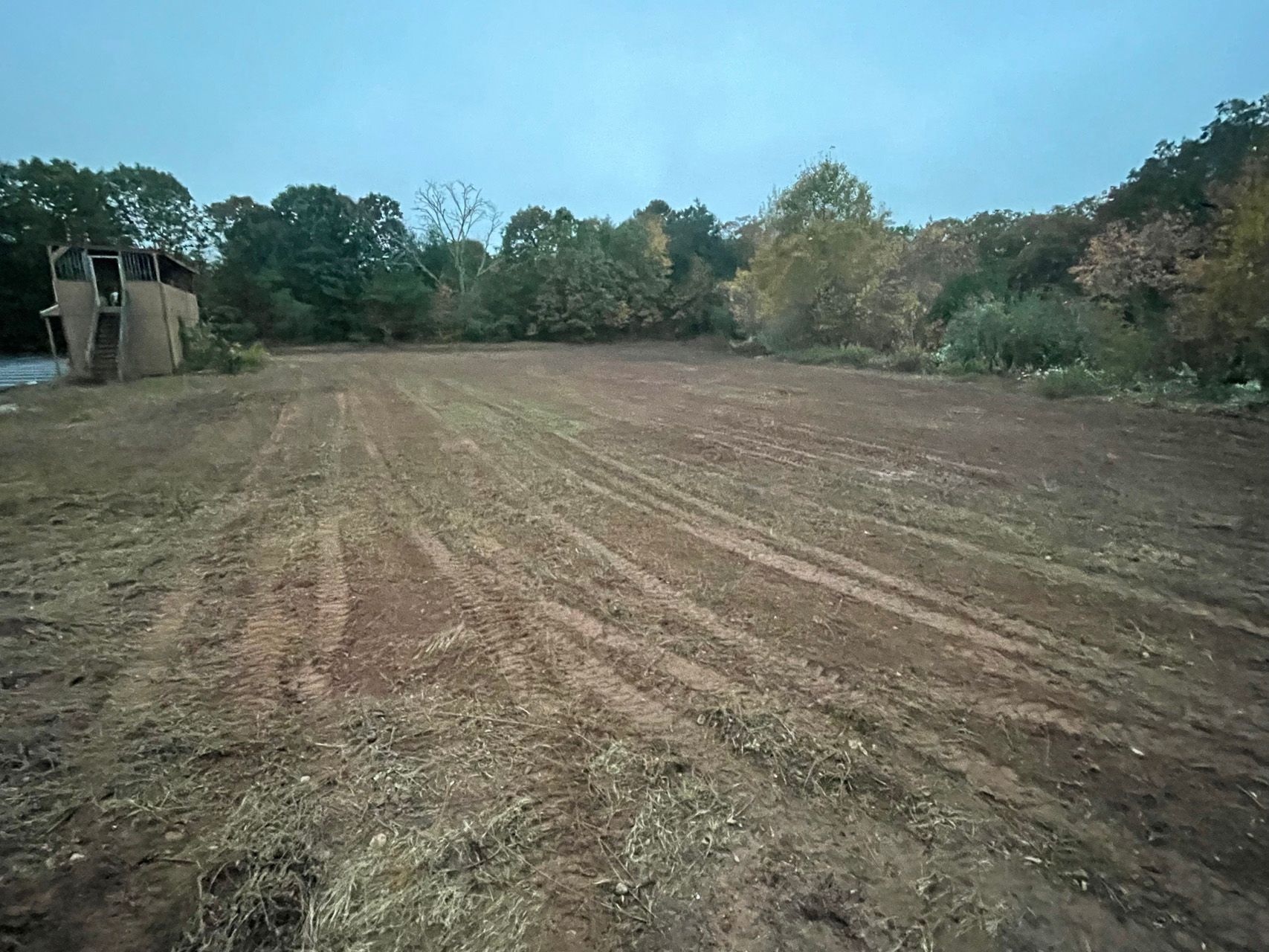 Recently plowed dirt field bordered by trees under a cloudy sky, with a small shed on the left.
