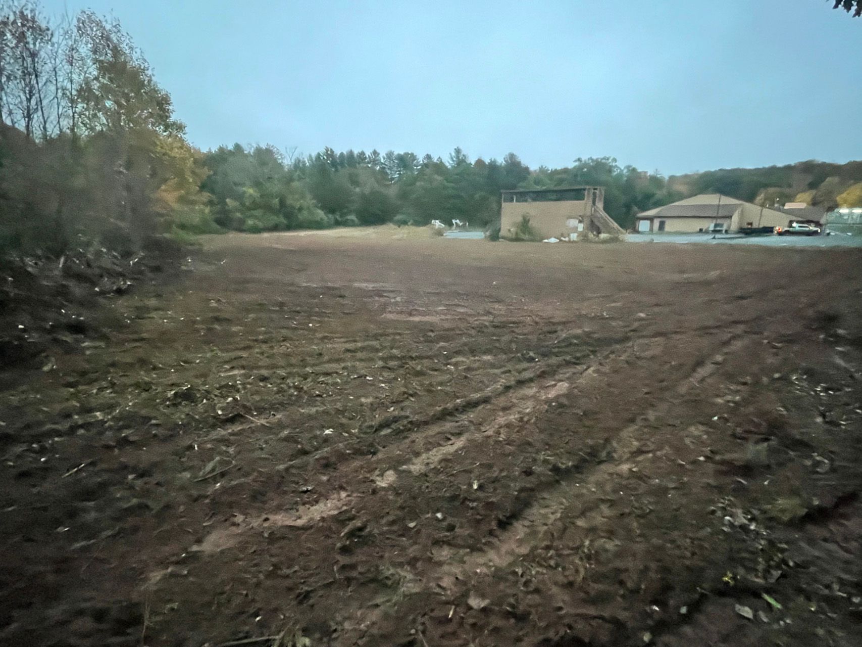 Freshly graded dirt lot beside a tree line, with a small house and parked vehicles in the distance.