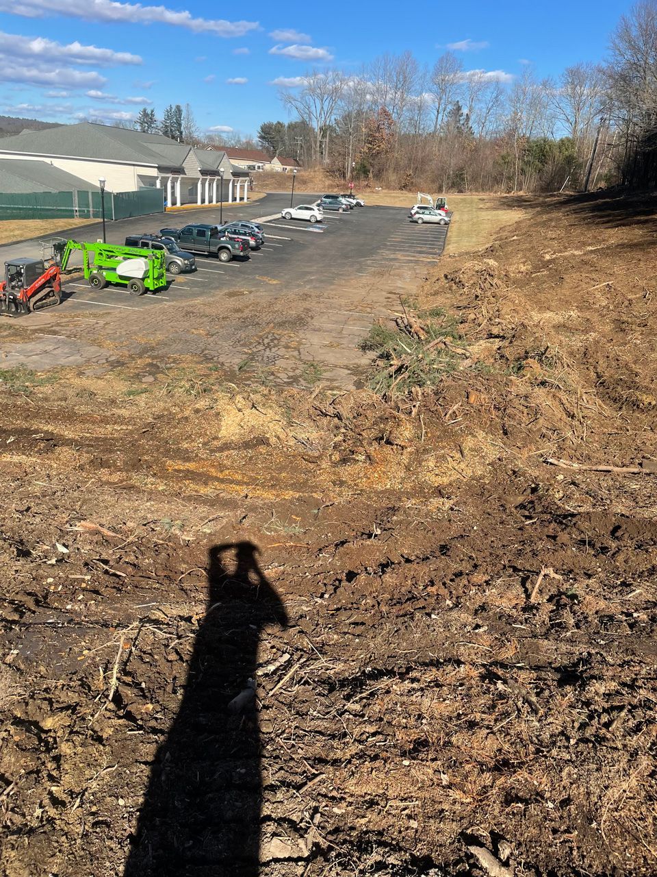 Dirt lot beside a greenhouse, with parked equipment under a clear blue sky and a long shadow in the foreground