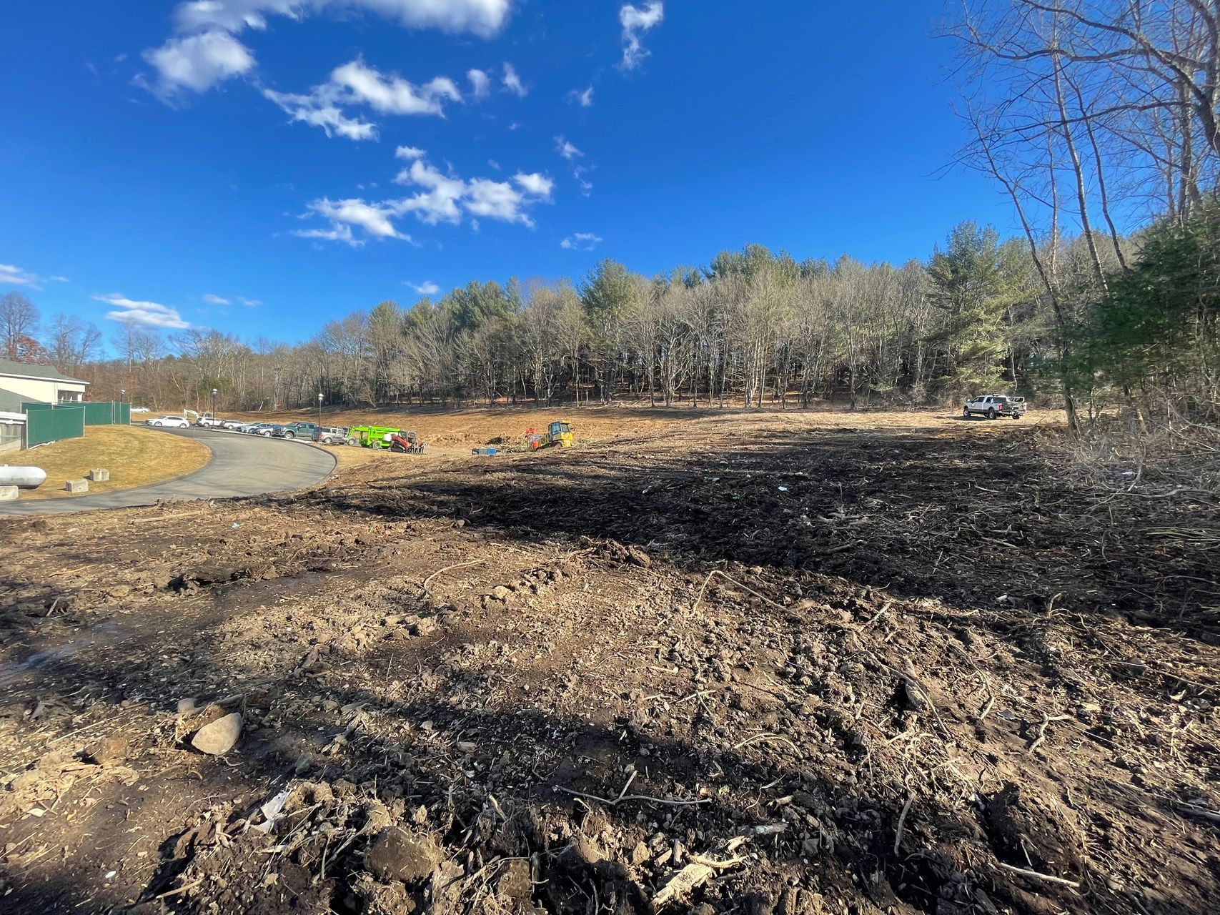 Cleared dirt lot under blue sky, with a tree line and a few parked vehicles in the distance
