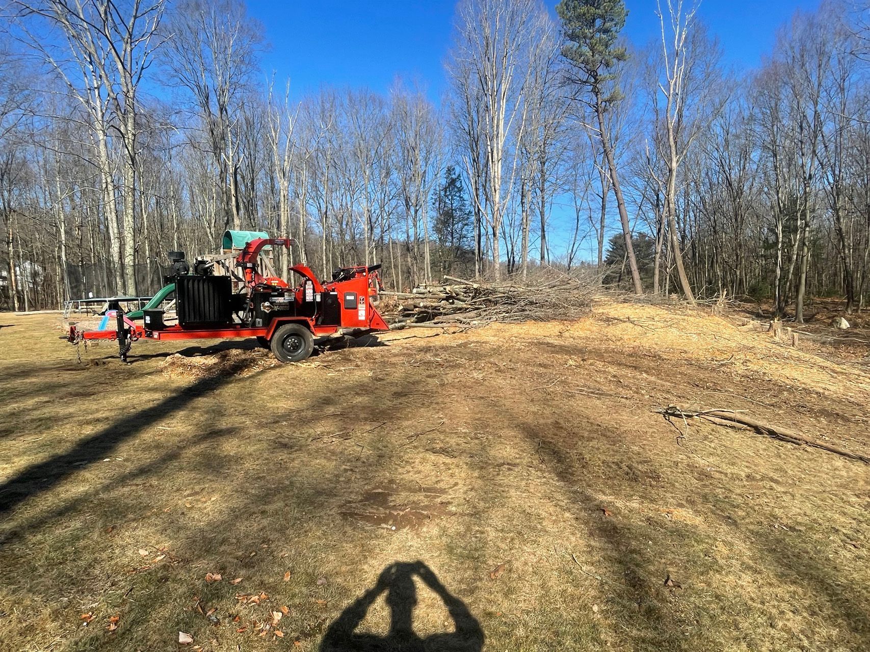 Red wood chipper beside a truck in a sunny wooded clearing with leafless trees.