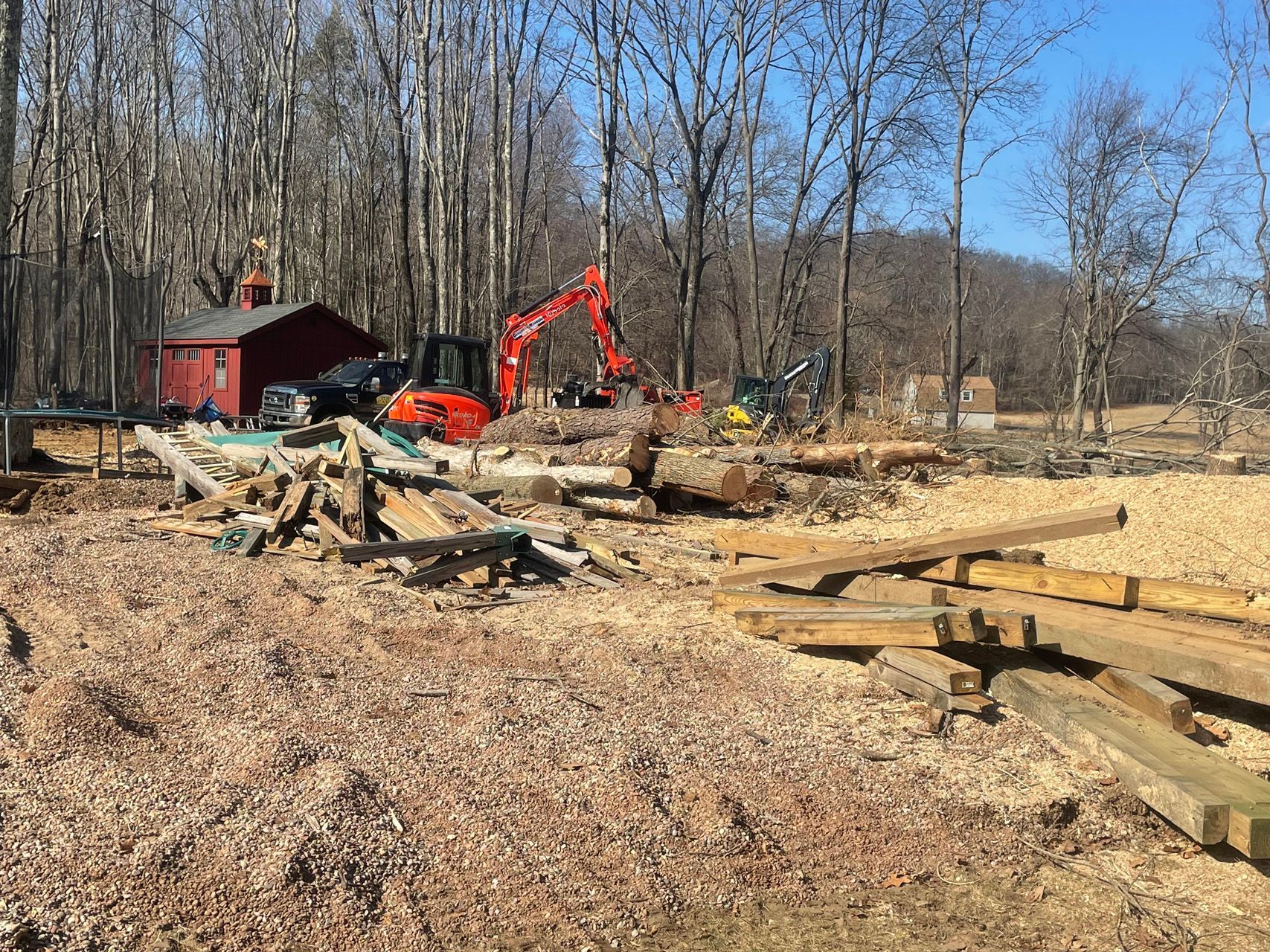 Red excavator and lumber in a cleared wooded area beside a red cabin on a sunny day