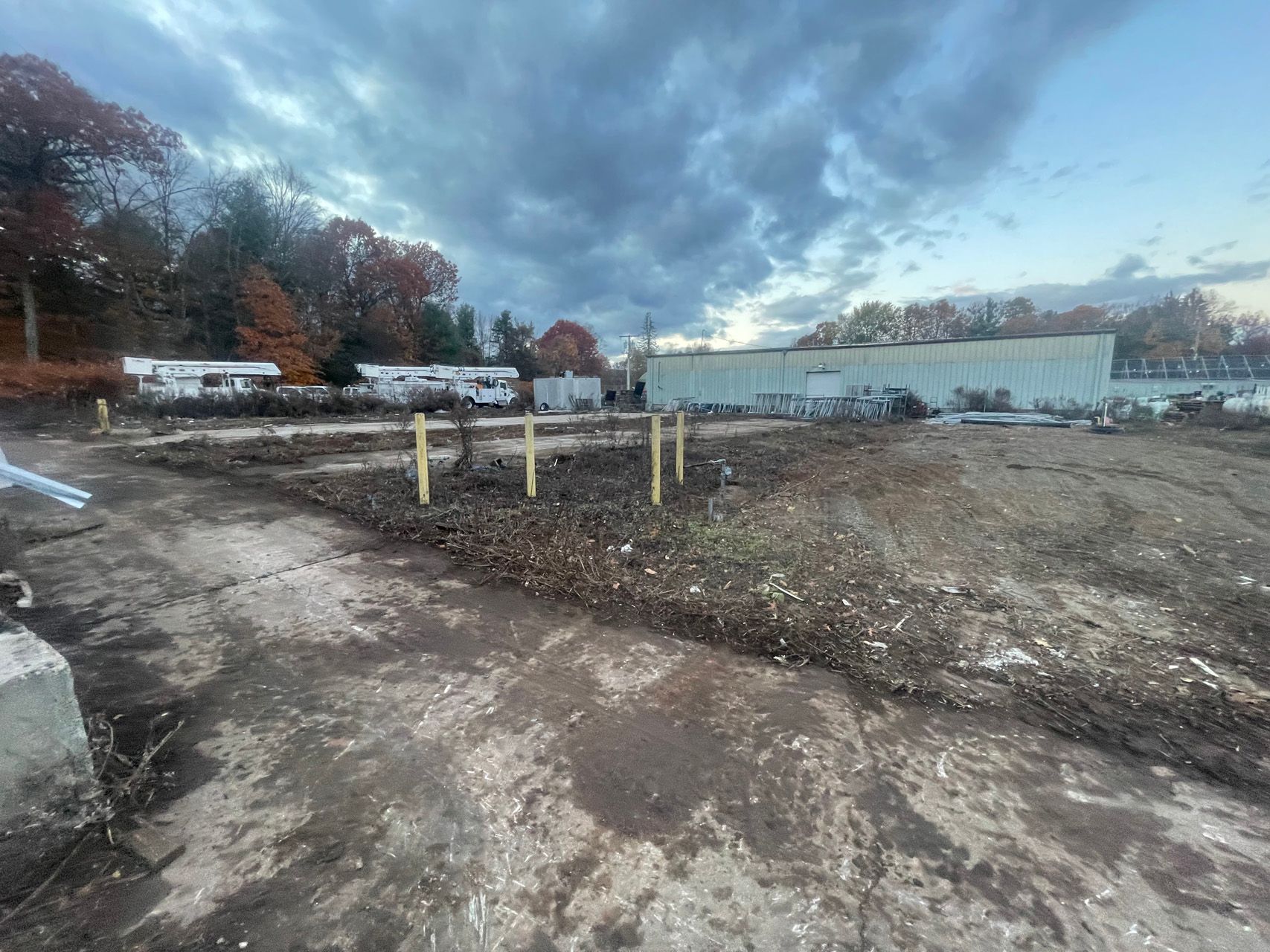 Dirt lot by a waterfront under cloudy sky, with parked vehicles and leafless trees in the background