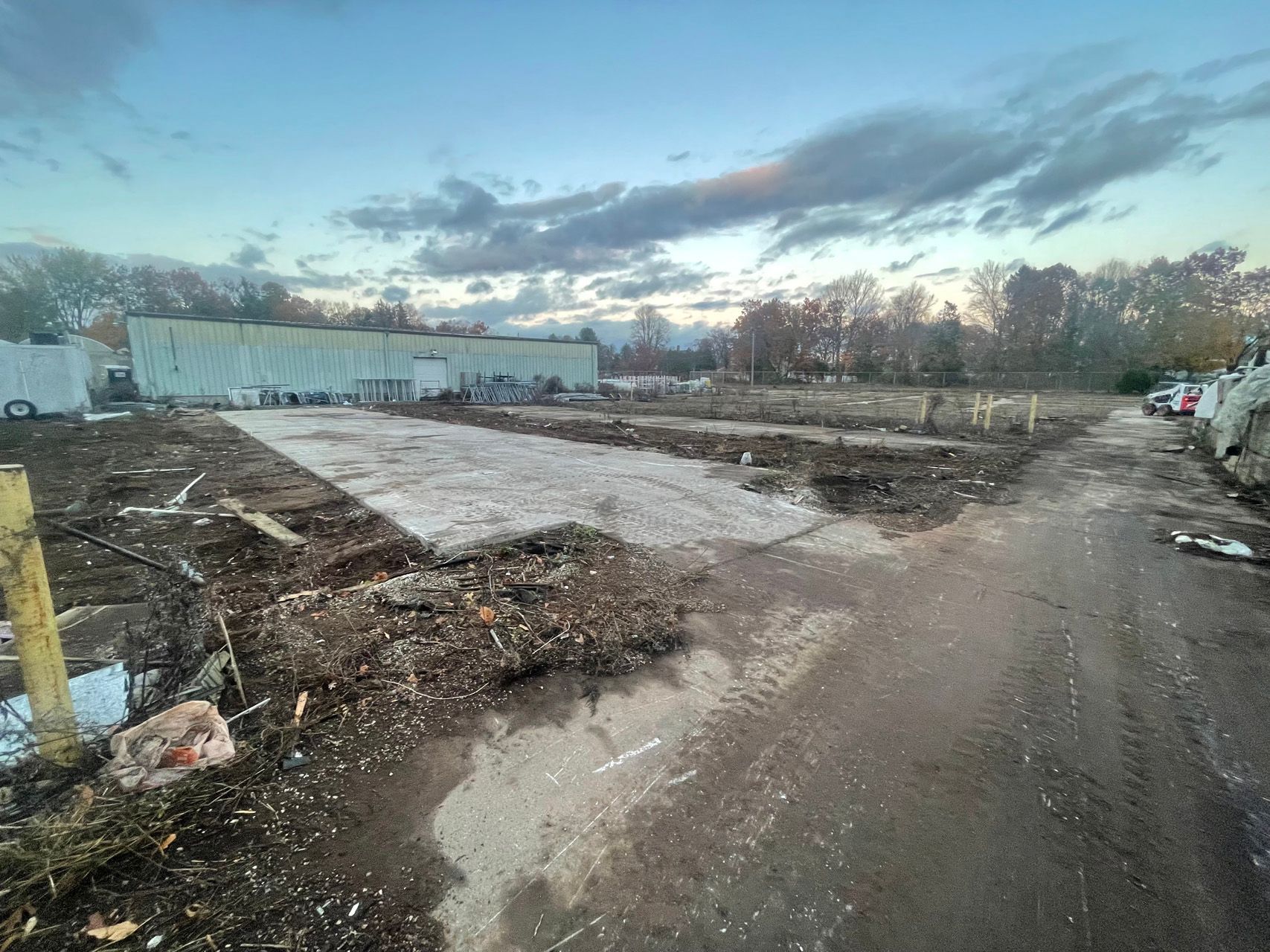 Muddy construction site at dusk with dirt piles, a paved path, and a large white building in the background