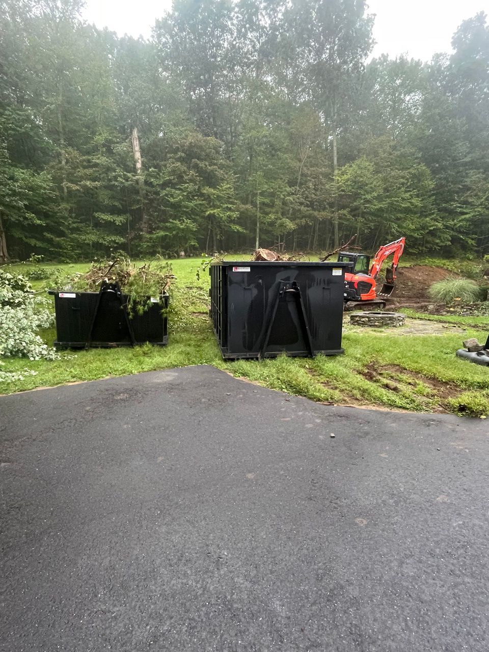 Black dumpsters by a wet driveway with an orange excavator in a foggy wooded area