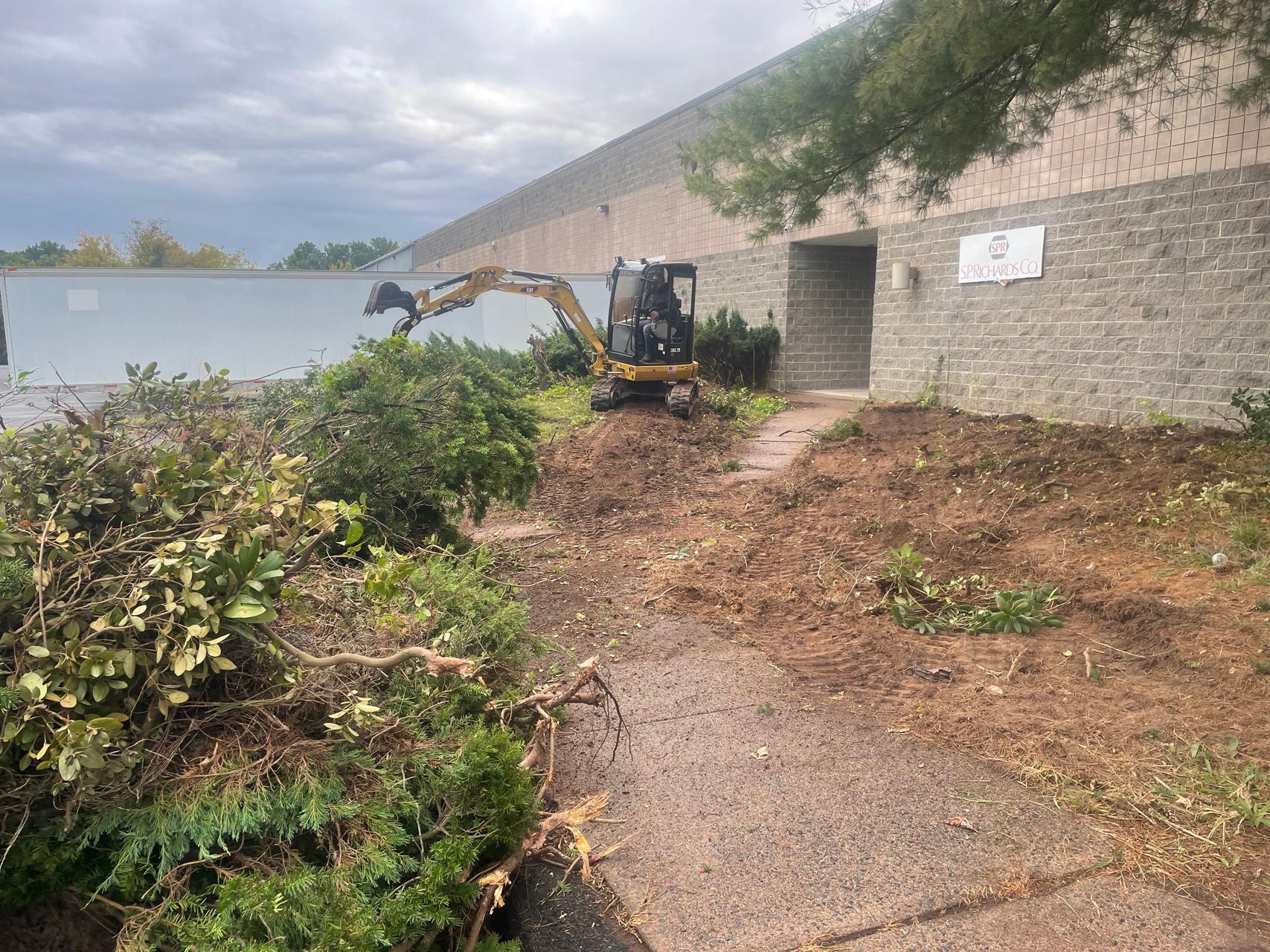 Small excavator clearing brush beside a stone building and walkway after storm damage
