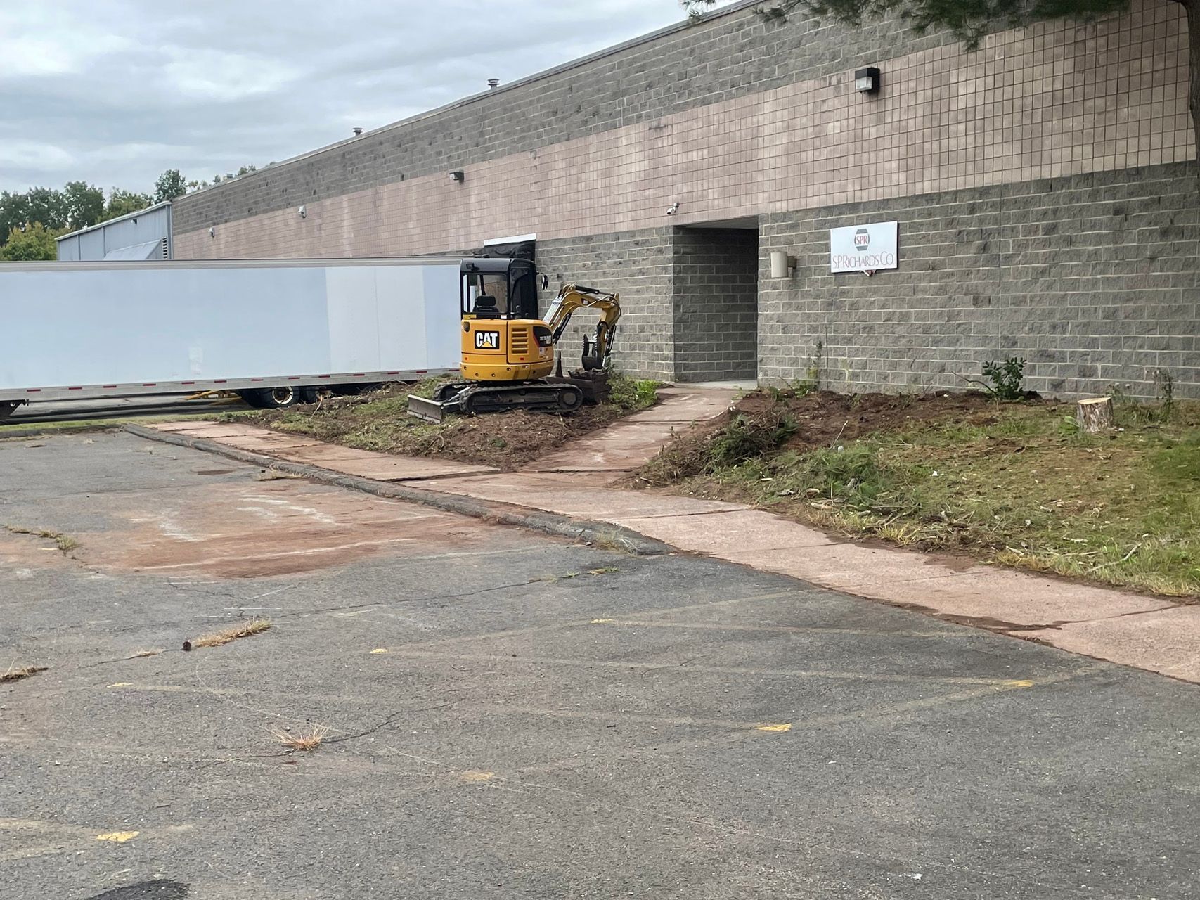 Yellow mini excavator beside a brick retaining wall, with a white truck trailer in an industrial lot.