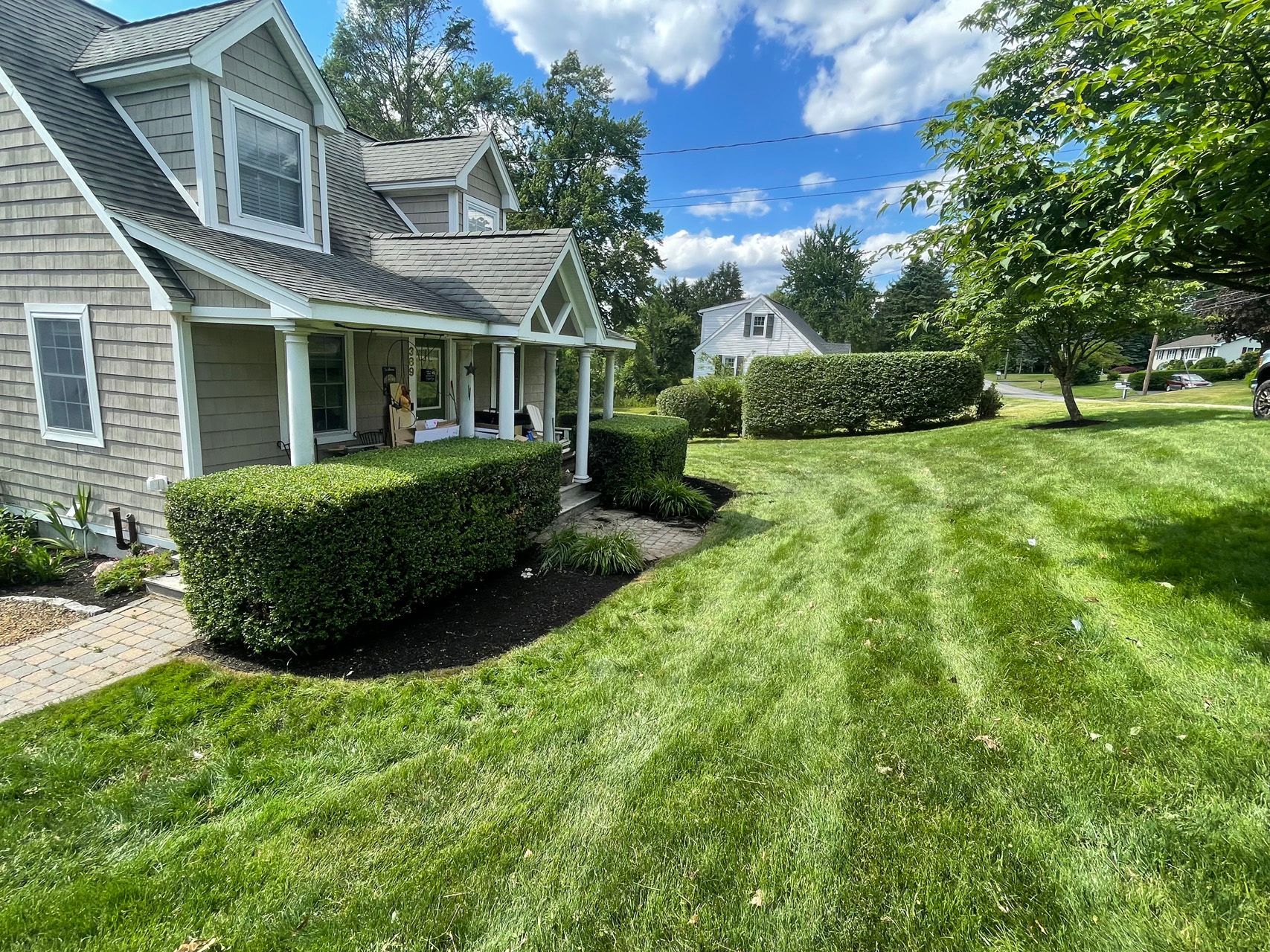 Suburban house with a front porch, manicured hedges, and a large green lawn under a blue sky.