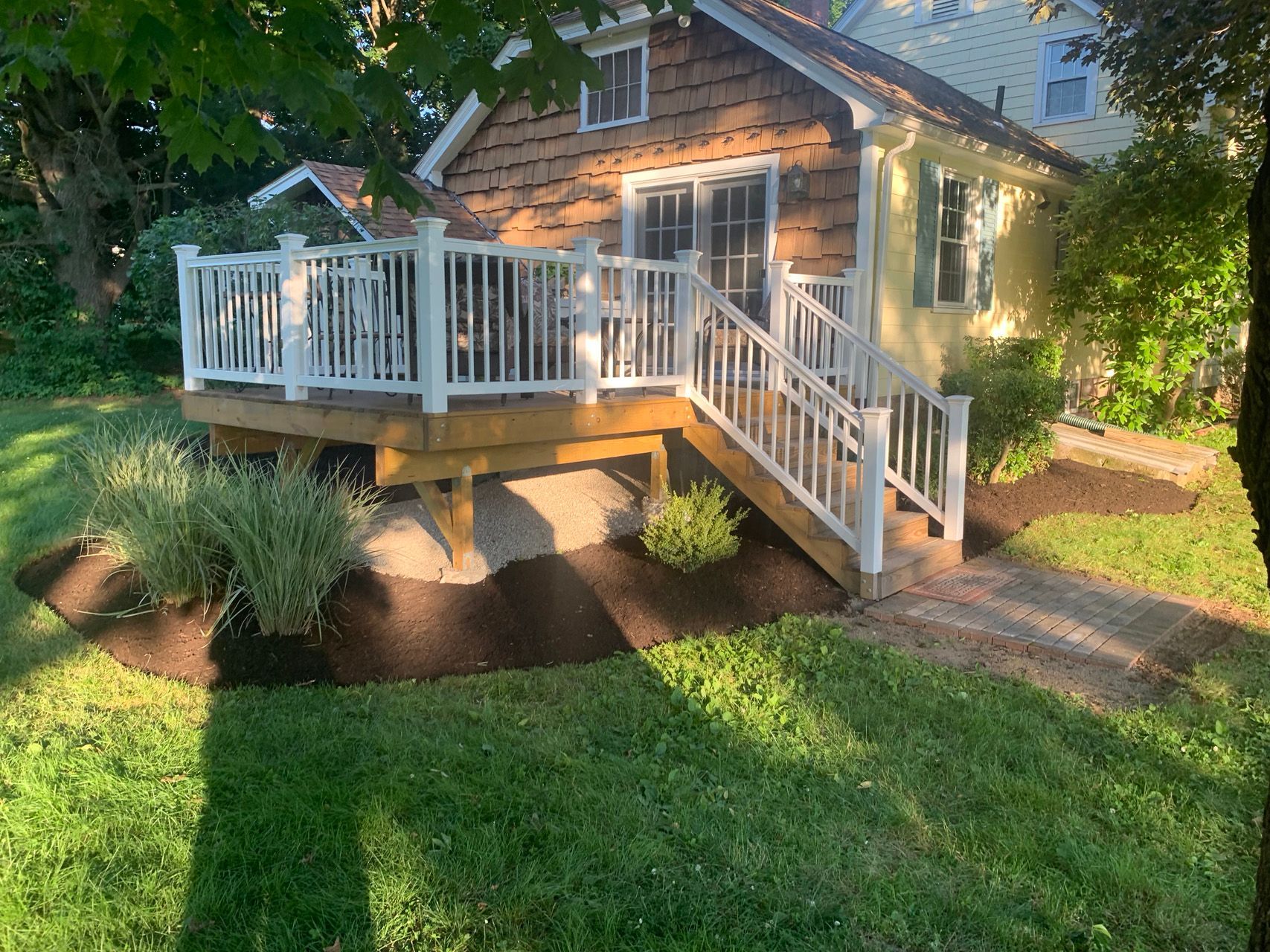 Small stone house with white porch and stairs, surrounded by green lawn and trees at sunset