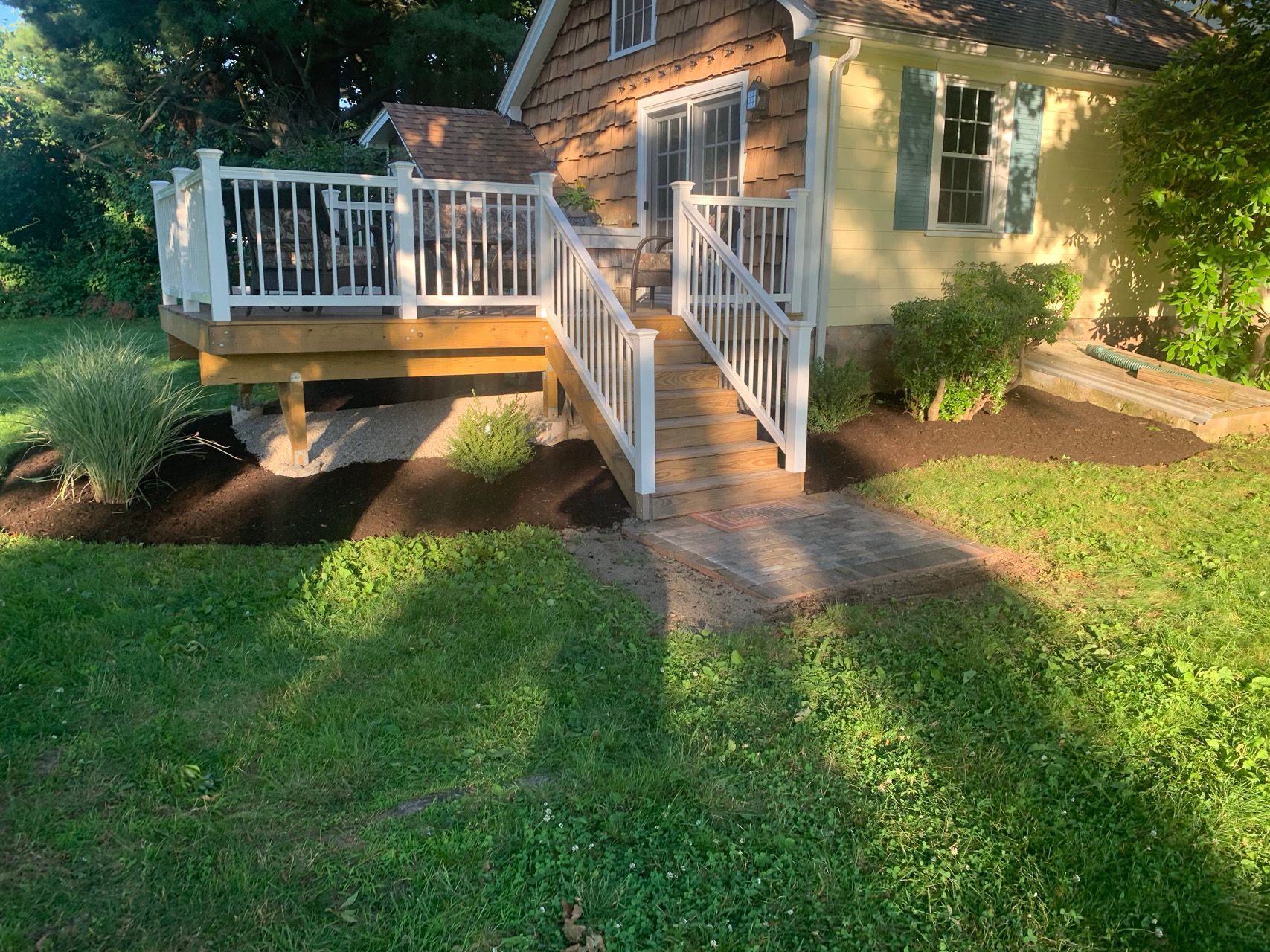 Small house with a wooden deck, white railing, stairs, and a grassy yard in warm sunlight.