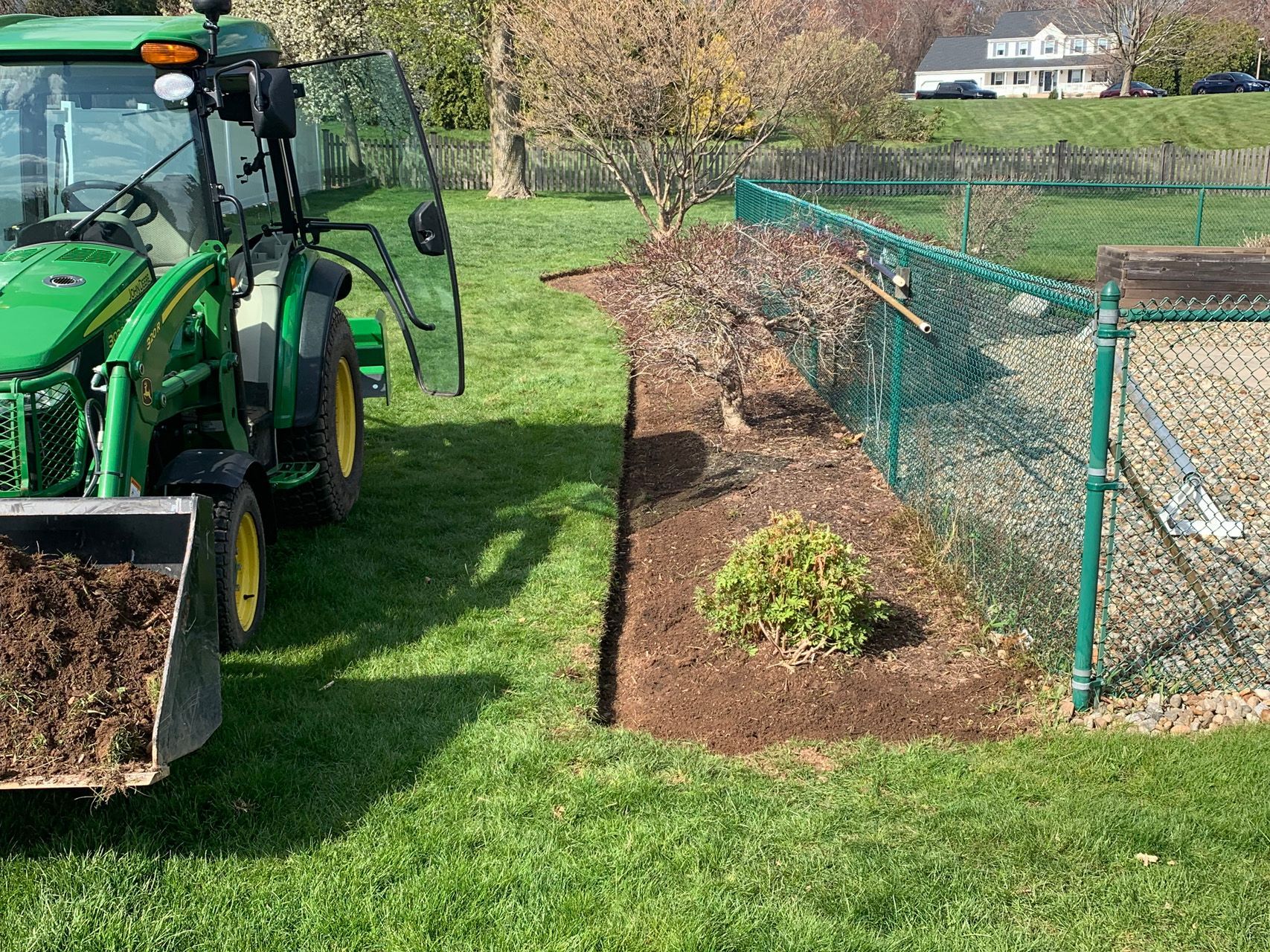 Green tractor beside a fenced garden bed being prepared with fresh mulch in a grassy yard