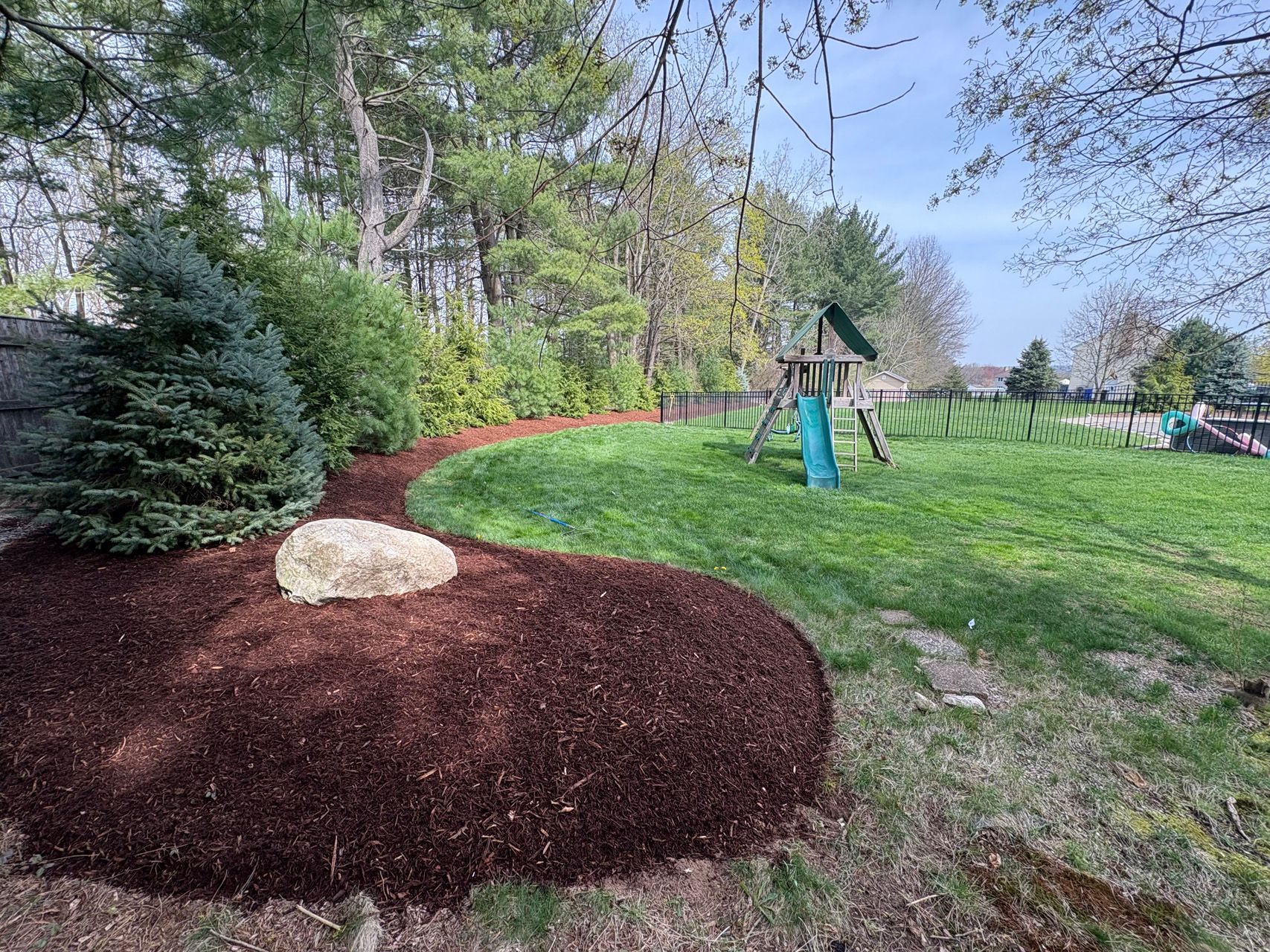 Landscaped park with a red mulch mound, rock, green lawn, and playground equipment under trees.