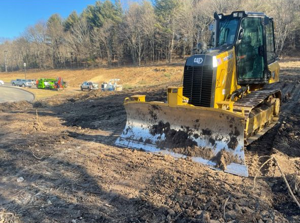 Yellow excavator working in a muddy field beside a water puddle, with trees in the background