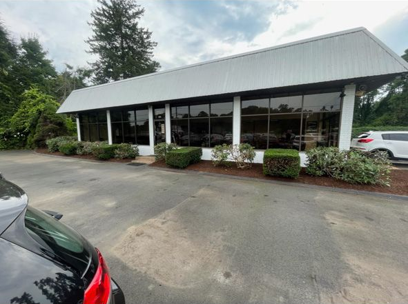 Covered outdoor building with white roof, surrounded by greenery, viewed from a parking area with a car in front